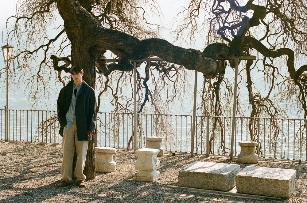 Giovane uomo indossa abbigliamento casual e si appoggia a un albero senza foglie vicino a un fiume, con una ringhiera e alcuni basamenti di pietra.