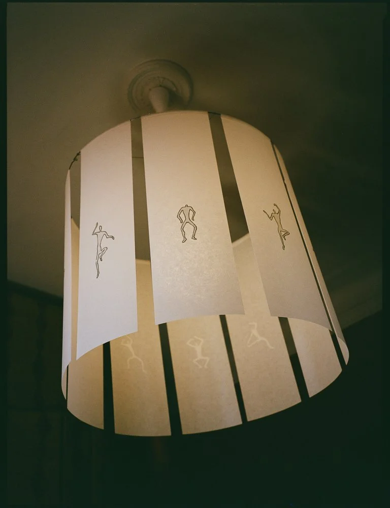 Lampada a sospensione con paralume decorato con disegni di figure umane stilizzate che sembrano ballare.