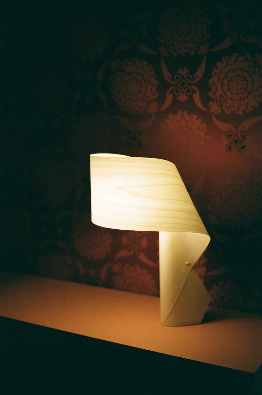 Lampada da tavolo accesa su una superficie con carta o legno chiaro, sfondo con carta da parati rossa scura con motivi decorativi