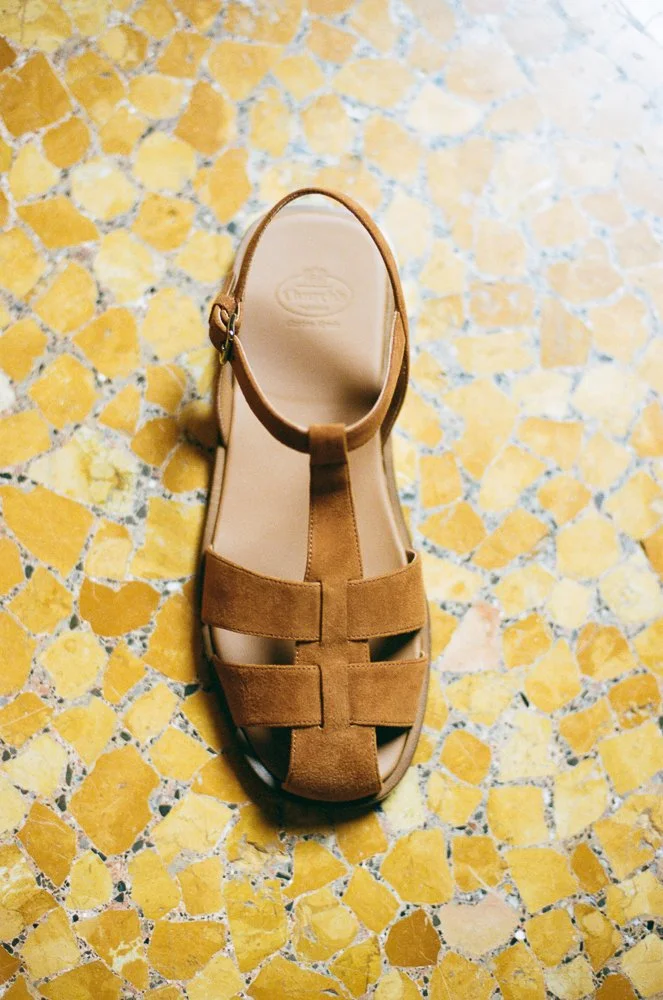 Sandalo marrone con cinturino anteriore e cinturino alla caviglia, posizionato su un pavimento di piastrelle gialle.