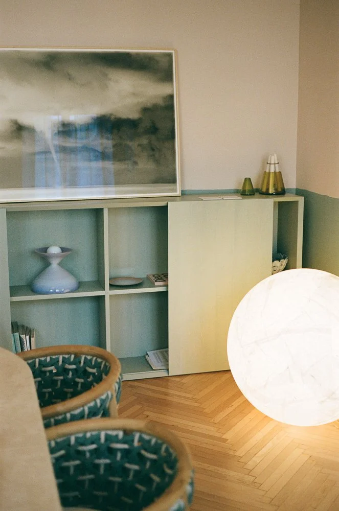 Interno con scaffale bianco, lampada di carta luminosa, sedie con rivestimento particolare, quadro con cielo nuvoloso.