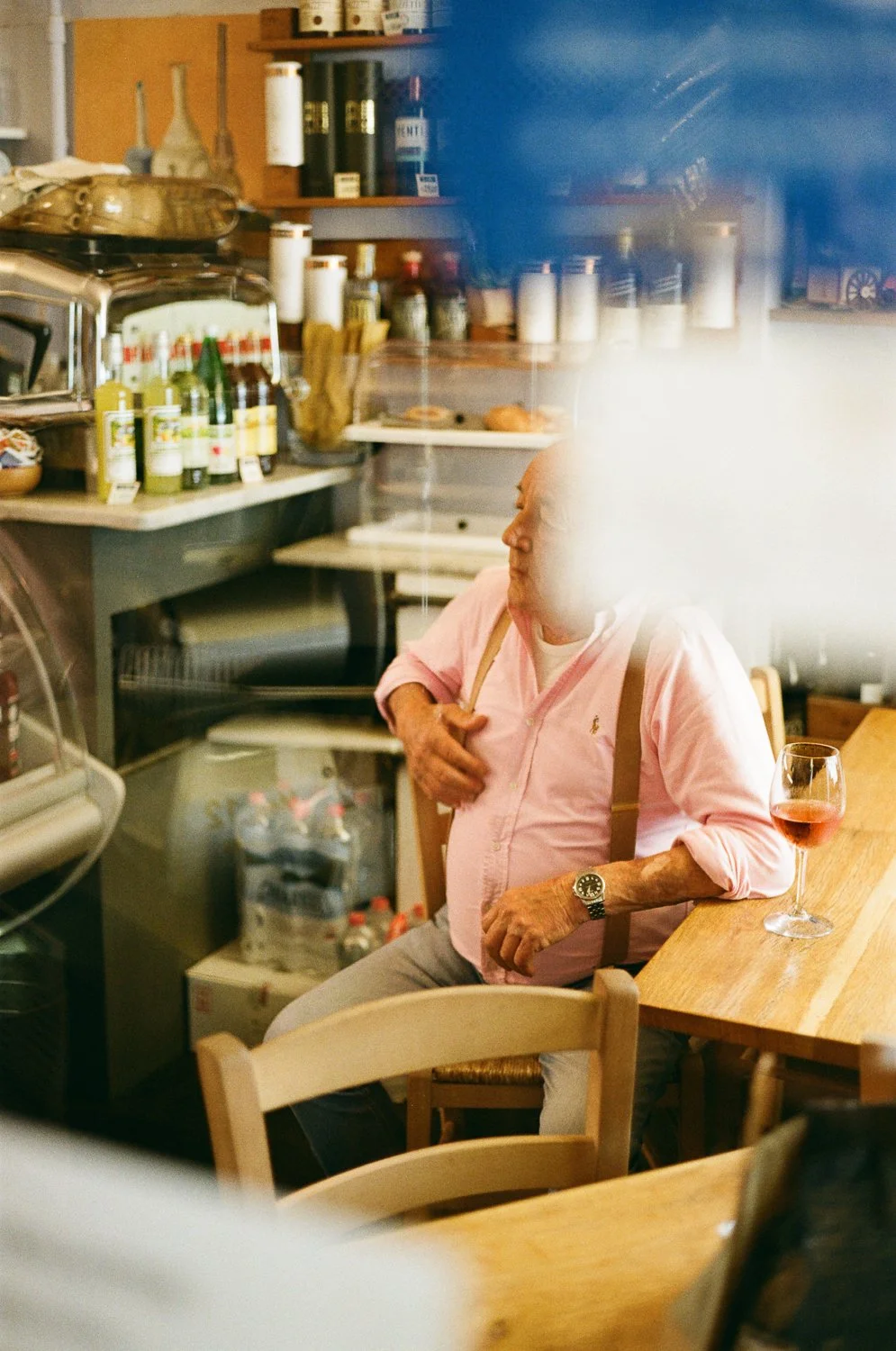 Un uomo anziano seduto in un bar con una tazza di vino rosato sul tavolo, indossa una camicia rosa chiaro e un orologio al polso. La scena è vista attraverso una finestra o un vetro con riflessi.