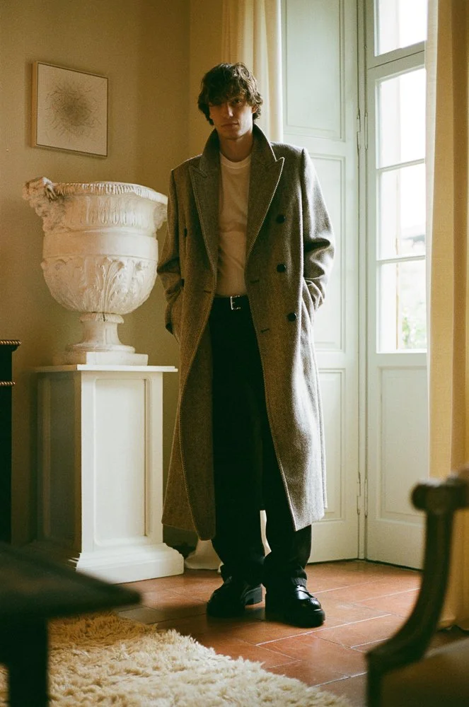 Una persona maschile con cappotto lungo grigio, camicia bianca e pantaloni neri, che si trova in una stanza luminosa con finestra e pareti beige.