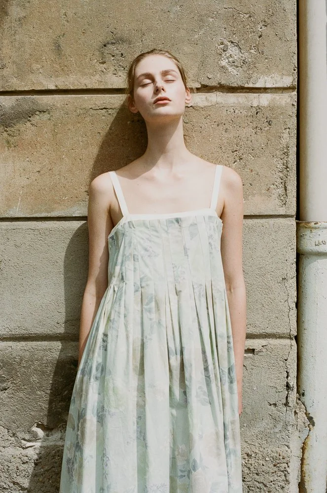 Ragazza con capelli castani chiusi, occhi chiusi, indossa un abito lungo con spalline sottili, appoggiata a un muro di pietra.