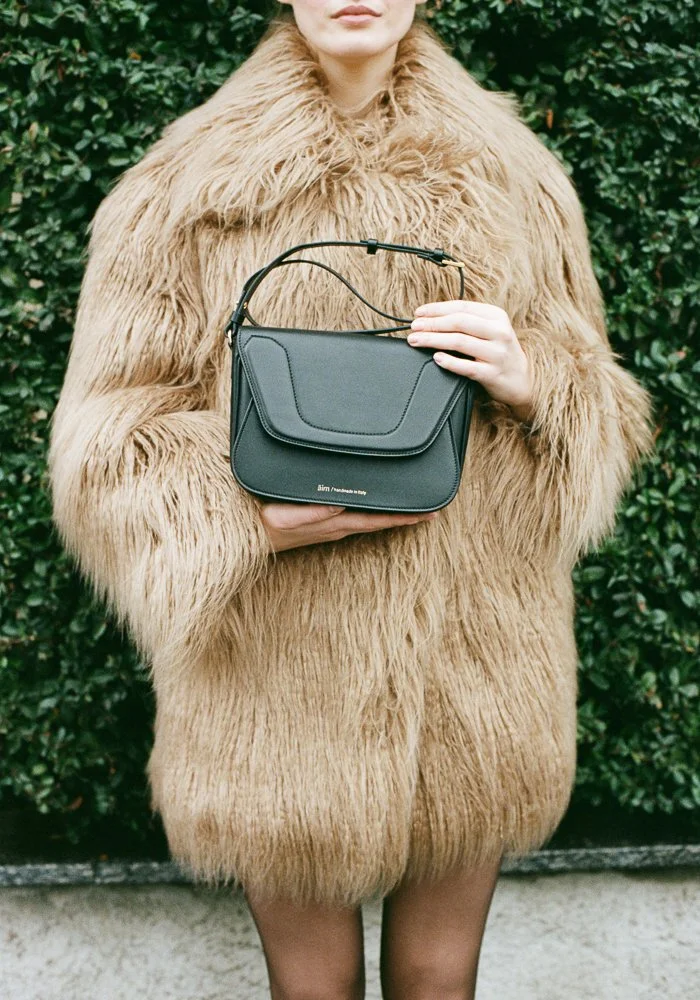 Donna indossa un cappotto di pelliccia beige e tiene in mano una borsa nera