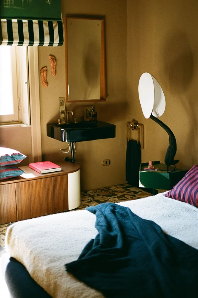 Camera da letto con letto coperto da una coperta beige, ci sono tessuti blu e a strisce, mobile in legno con libri e cuscini, specchio e lavabo nero sopra un mobile, lampada moderna accanto al letto.