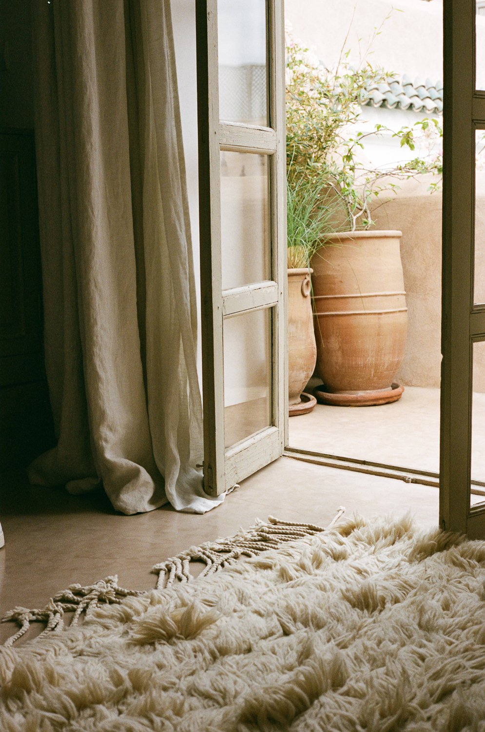 Porta di vetro aperta che dà su un balcone con grandi vasi in terracotta con piante verdi, vicino a tende chiare, sul pavimento di piastrelle. In primo piano un tappeto morbido e bianco con dettagli intrecciati.