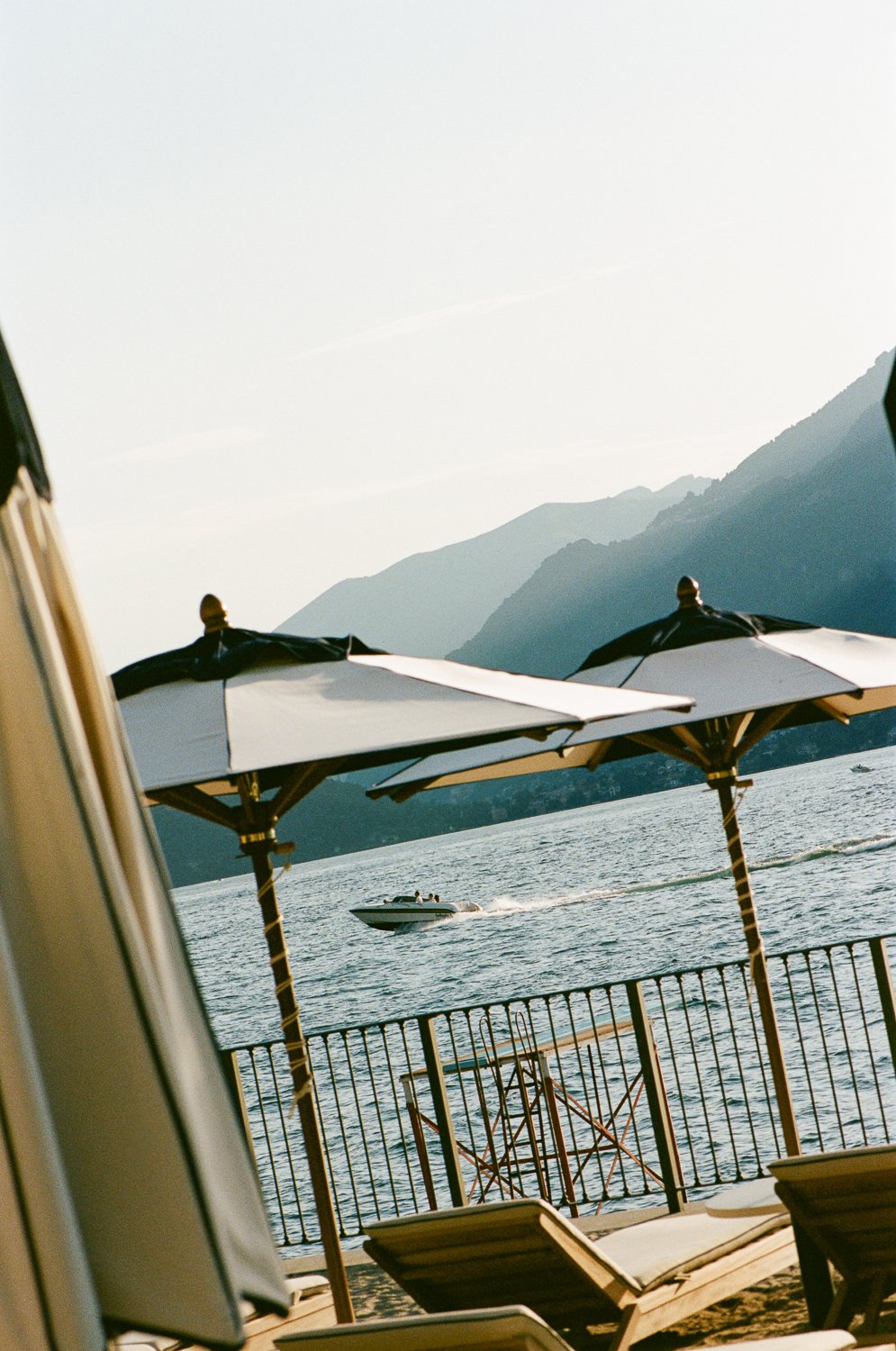 Veduta di un lago con montagne sullo sfondo, un motoscafo in movimento e ombrelloni sulla spiaggia.
