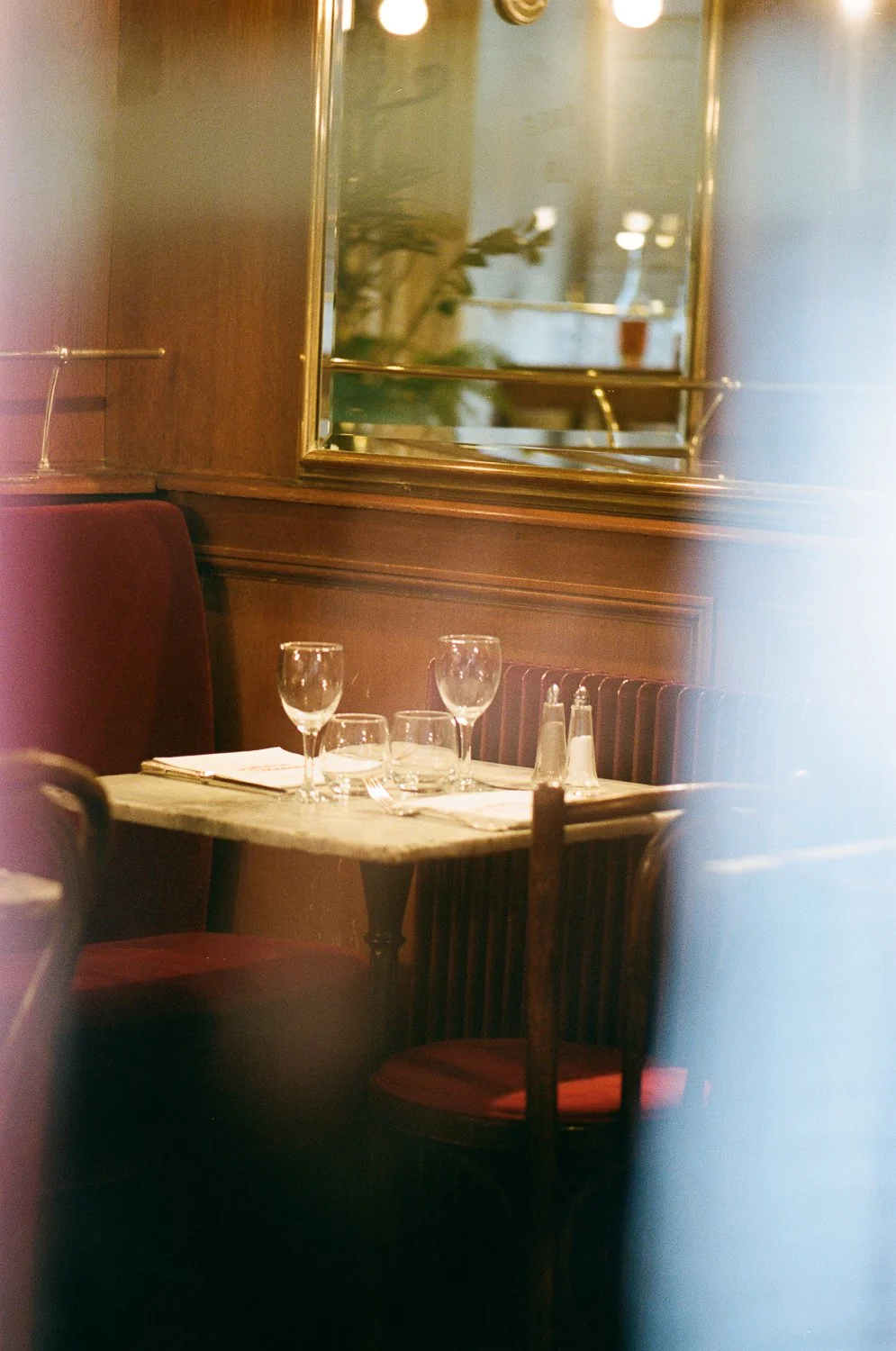 Tovaglia apparecchiata con due bicchieri da vino, bicchieri d'acqua e posate, in un ristorante con sedie di velluto rosso e pareti di legno.