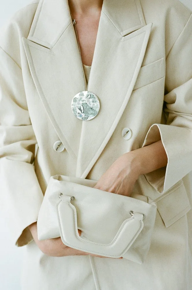 Donna in abito beige con borsa coordinata, indossa una giacca con ampi risvolti e una grande spilla decorativa, tiene una borsa bianca con manici corti.