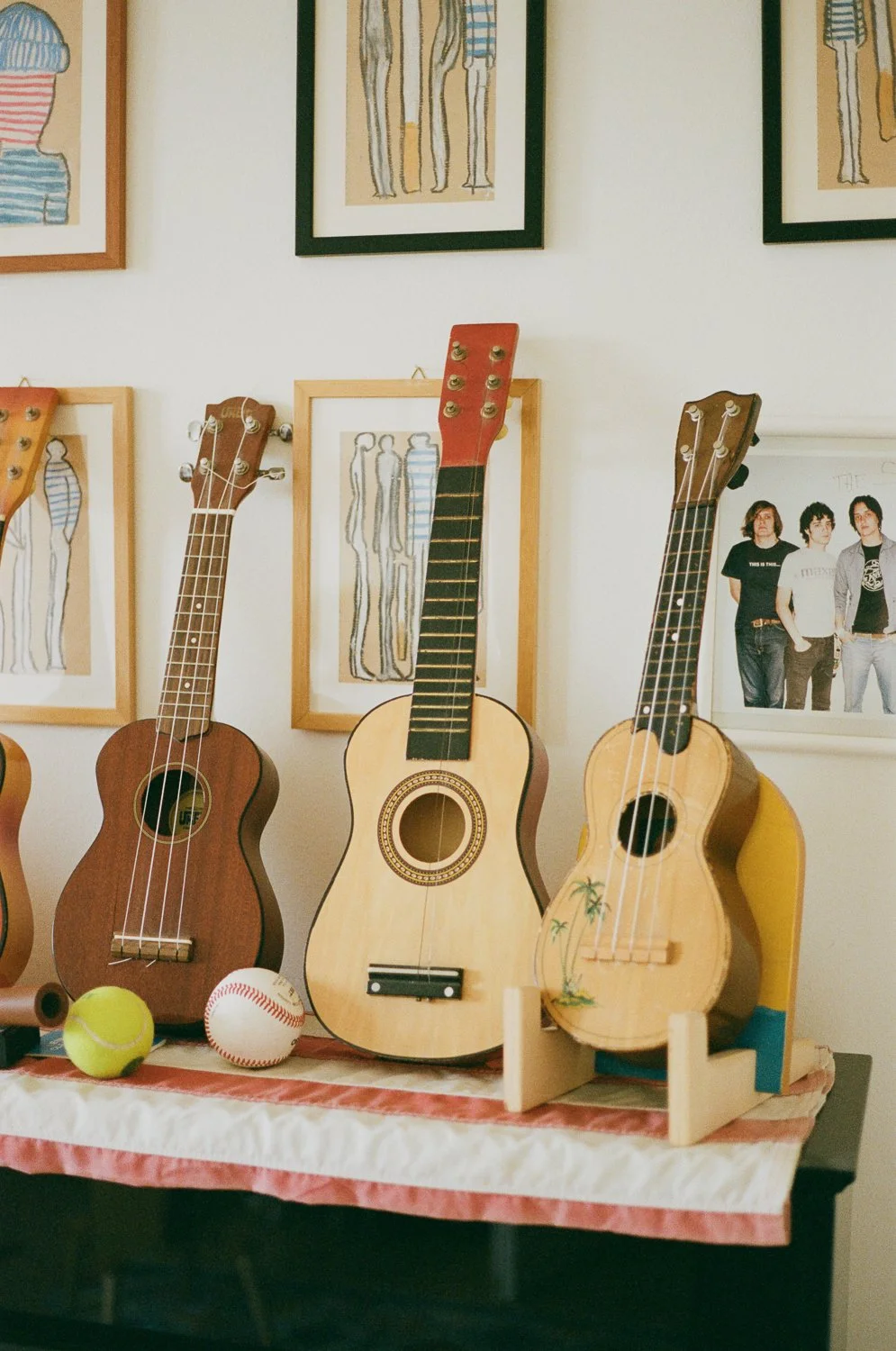 Tre piccole chitarre acustiche di legno posizionate su un tavolo con palline da tennis e da baseball davanti. Sullo sfondo, quadri con disegni stilizzati di figure umane e una foto di tre giovani.