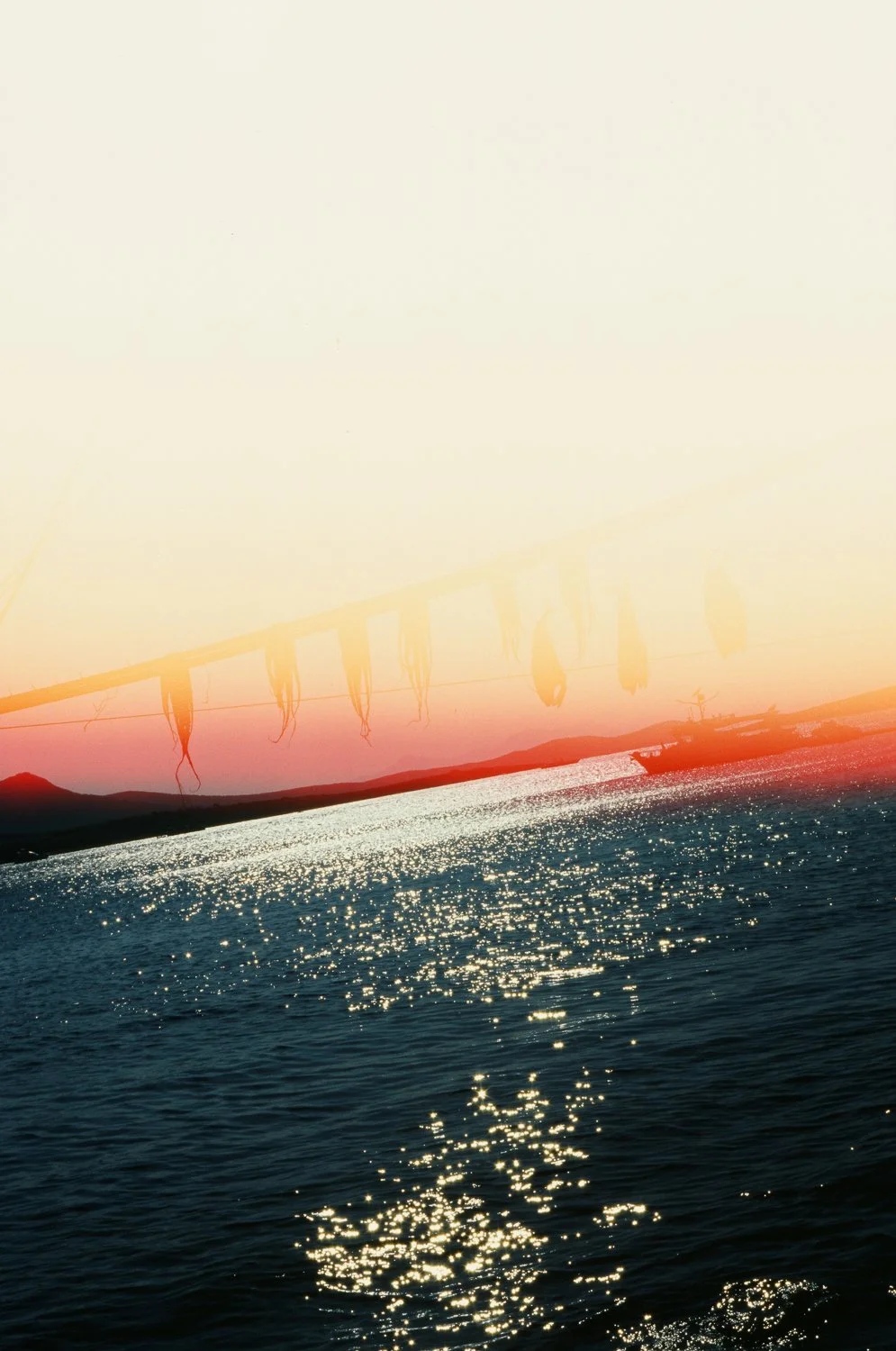 Il mare con il sole che riflette sull'acqua, al tramonto, con qualche montagna sullo sfondo e delle barche in lontananza.