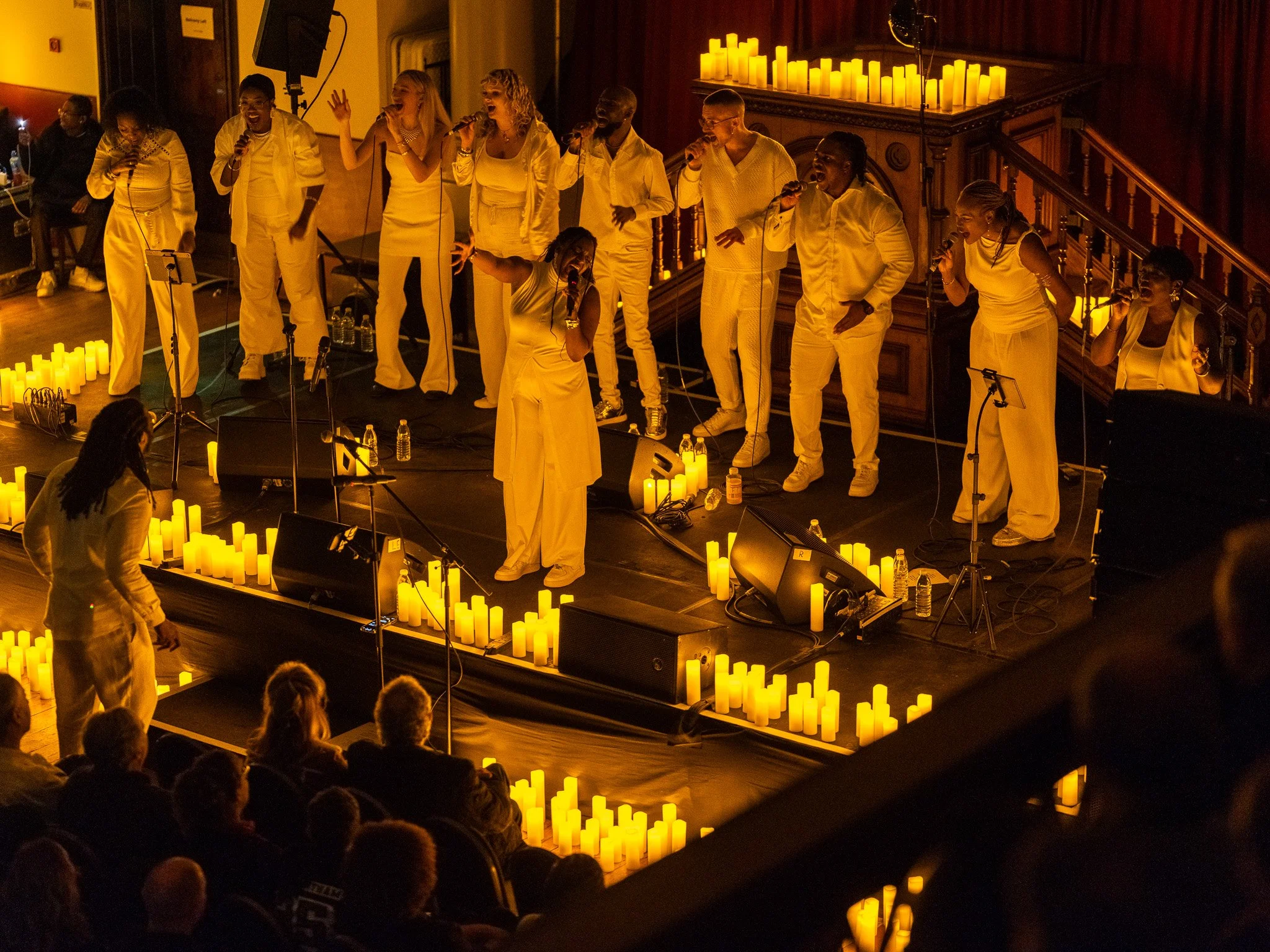 A choir of people dressed in white performing on a stage decorated with candles, with an audience watching.