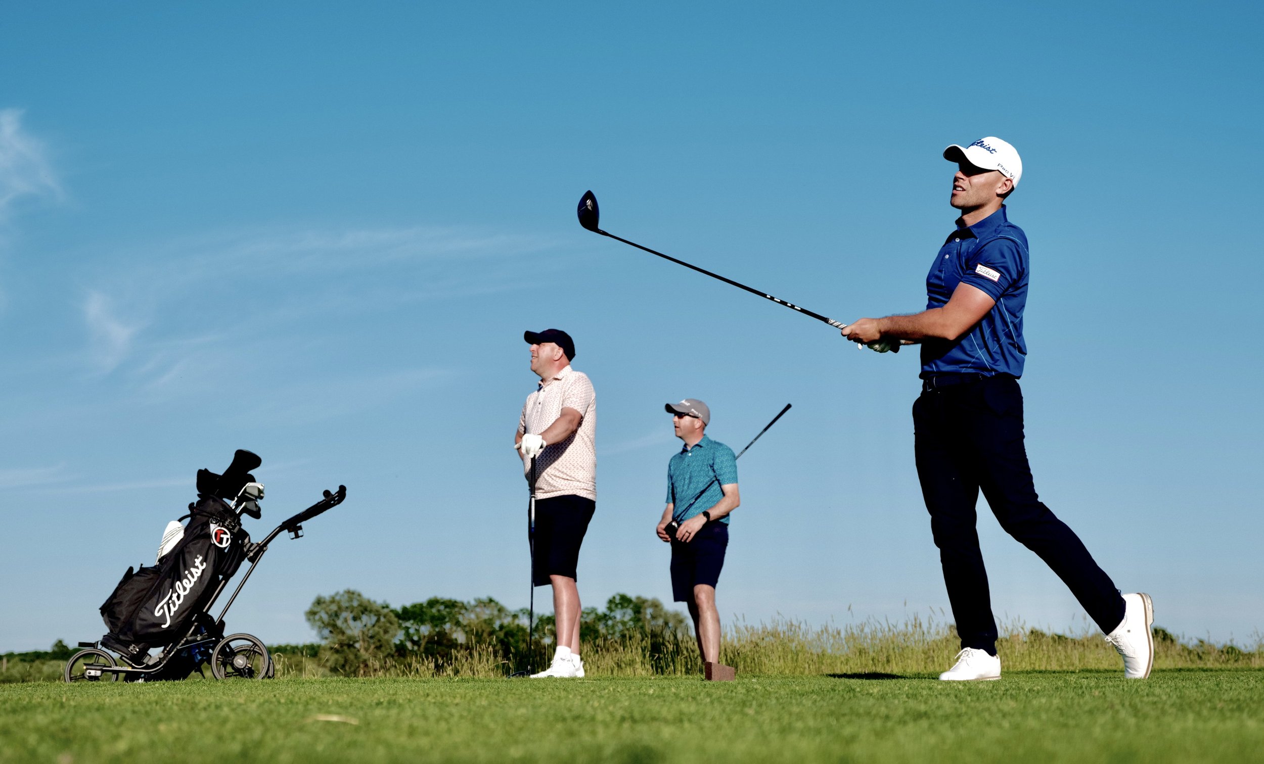 Three men playing golf on a sunny day with a game trolley nearby.