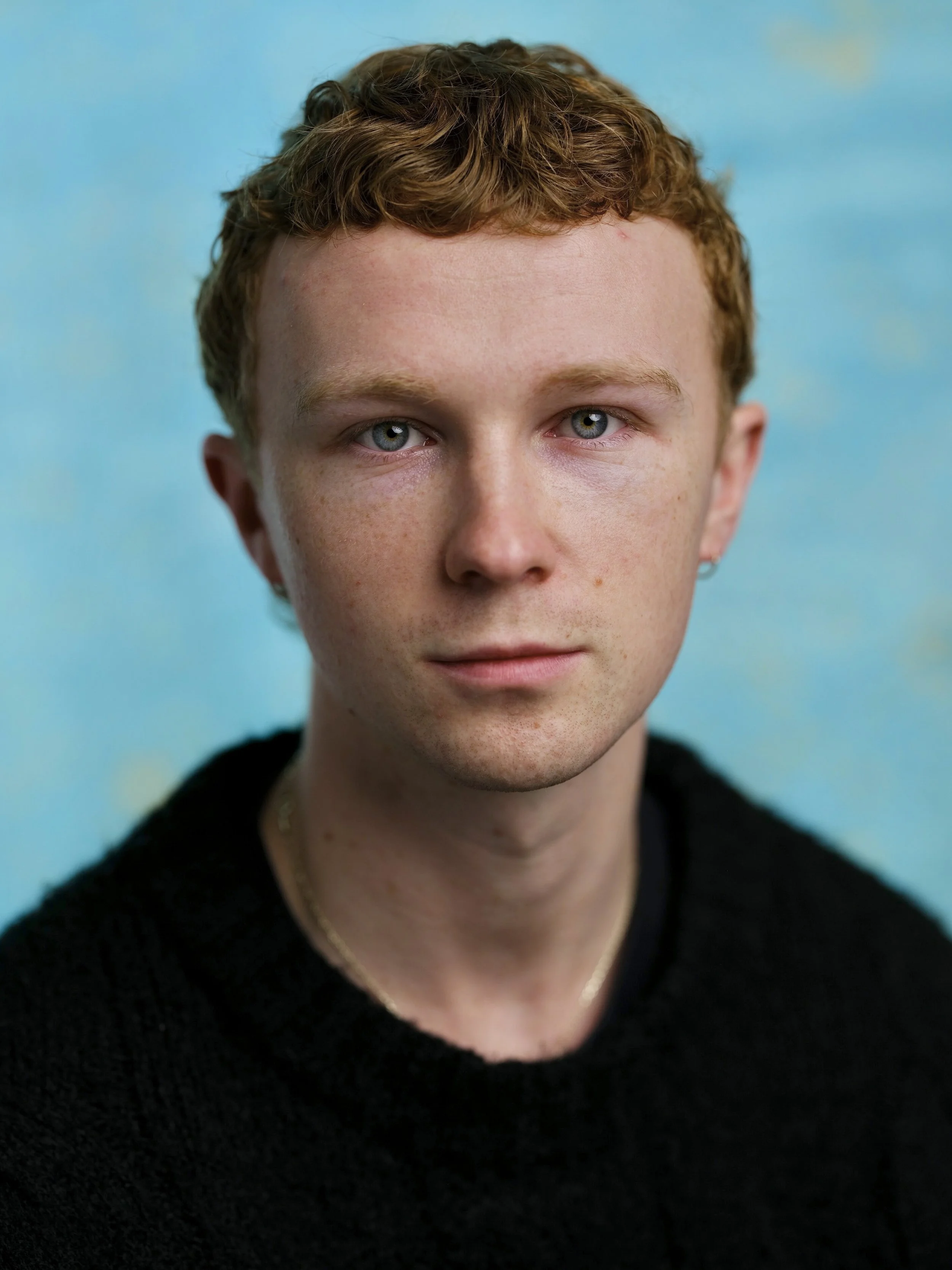 A young man with short, curly red hair and blue eyes, wearing a black sweater and gold chain, against a blue sky background.