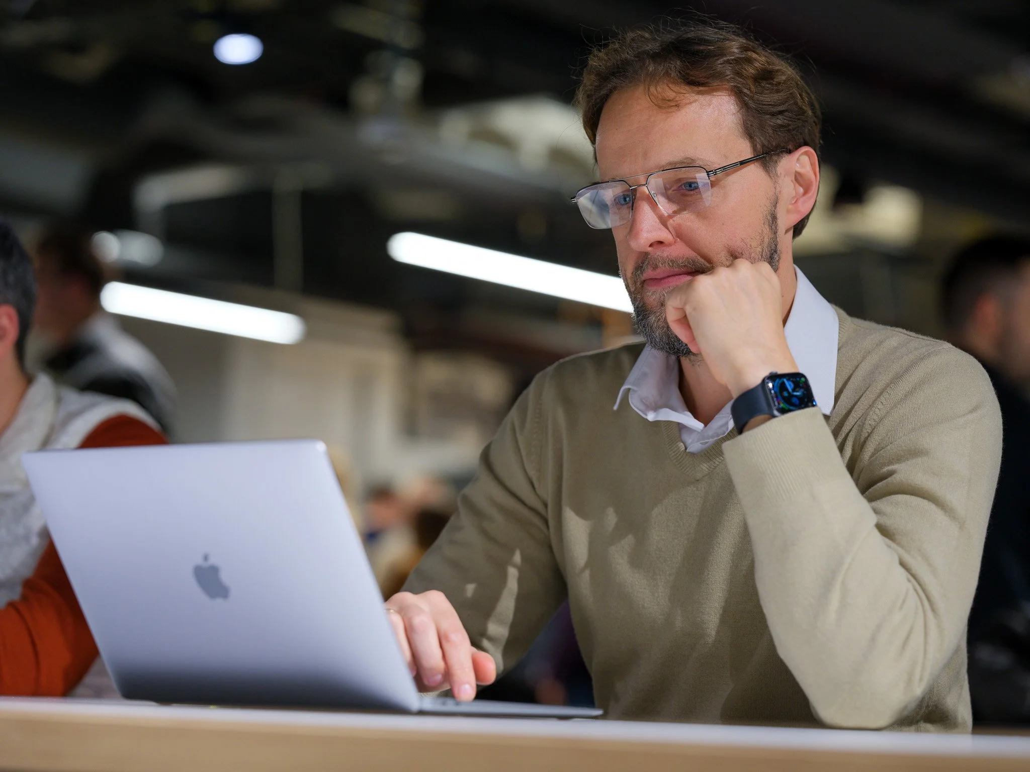 A man with glasses and a beard working on a silver MacBook at a table, wearing a beige sweater over a white shirt, in a shared workspace or cafe.