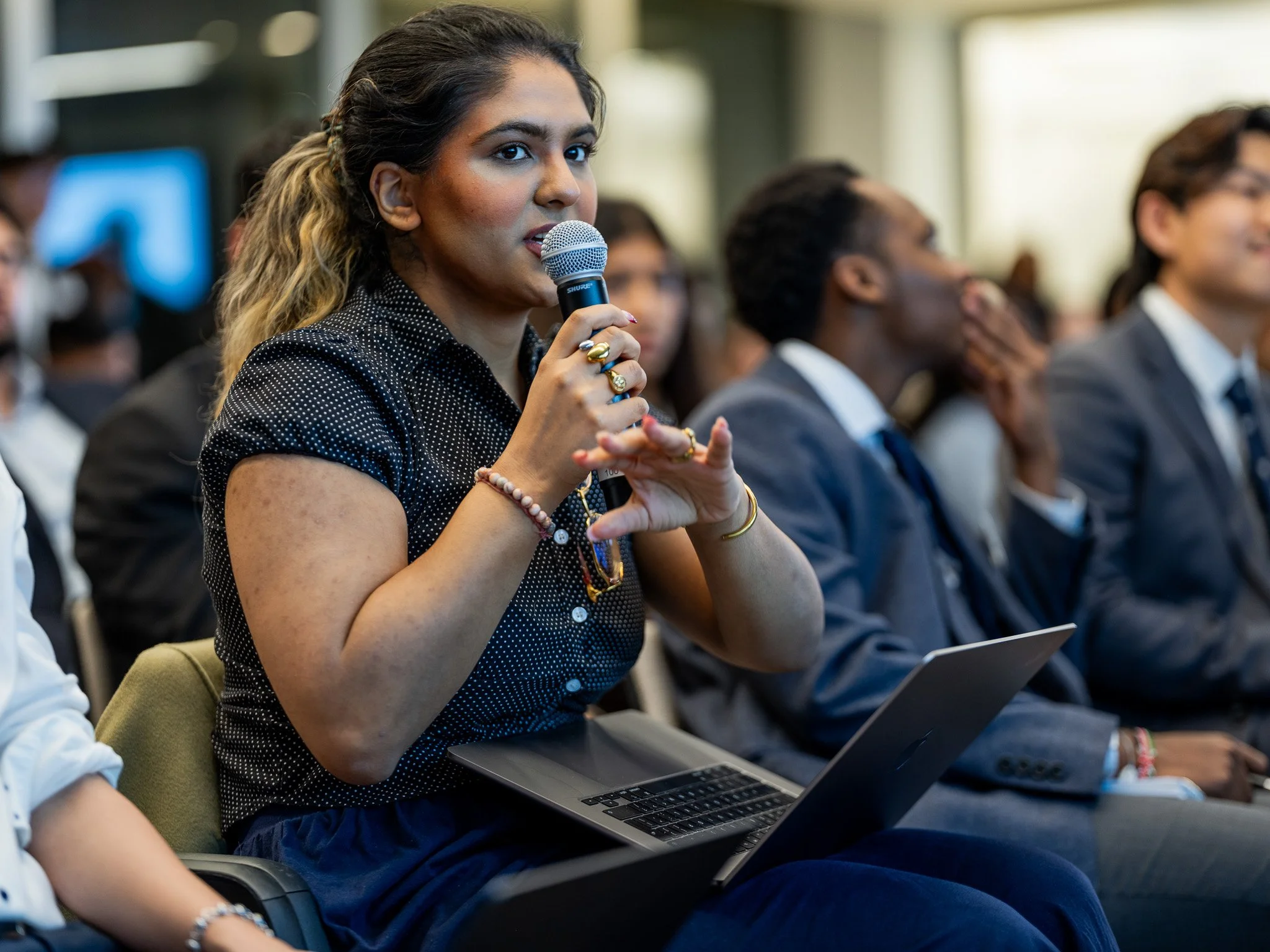 A diverse group of people attending a conference, with a woman in the foreground holding a microphone and a laptop on her lap, speaking or asking a question.