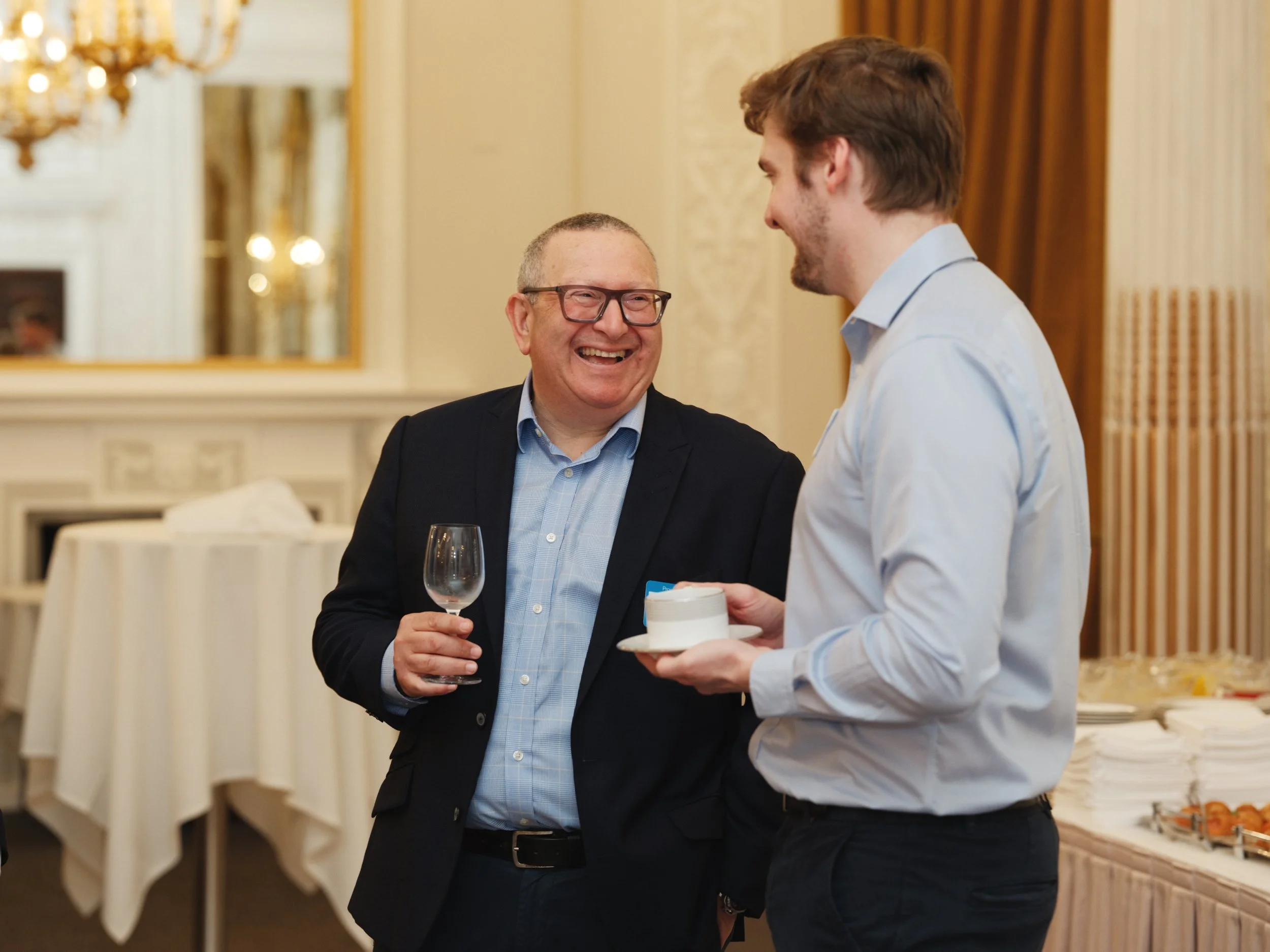 Two men talking at a social event in an elegant room; one holding a glass of wine and the other a small plate with a dessert.