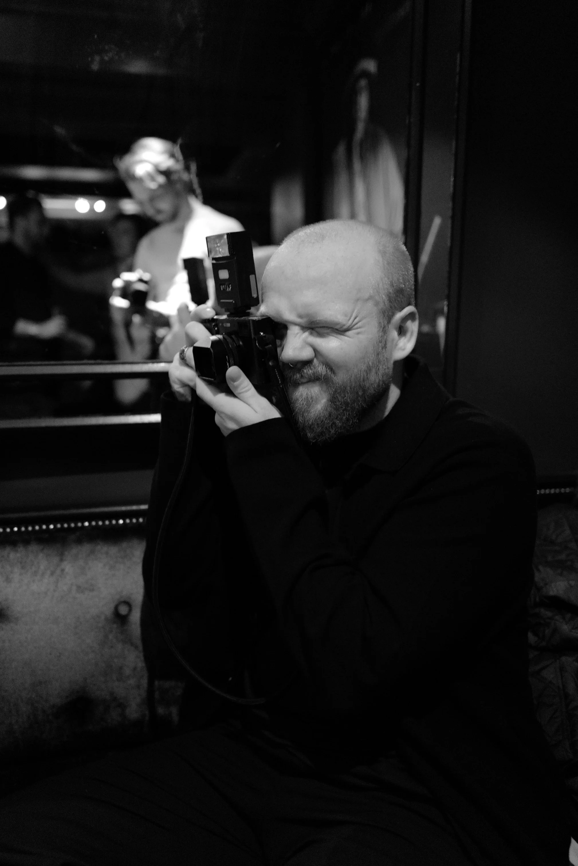 A man with a beard and bald head taking a photograph with a camera, sitting indoors near a mirror, with other people and artwork visible in the background.