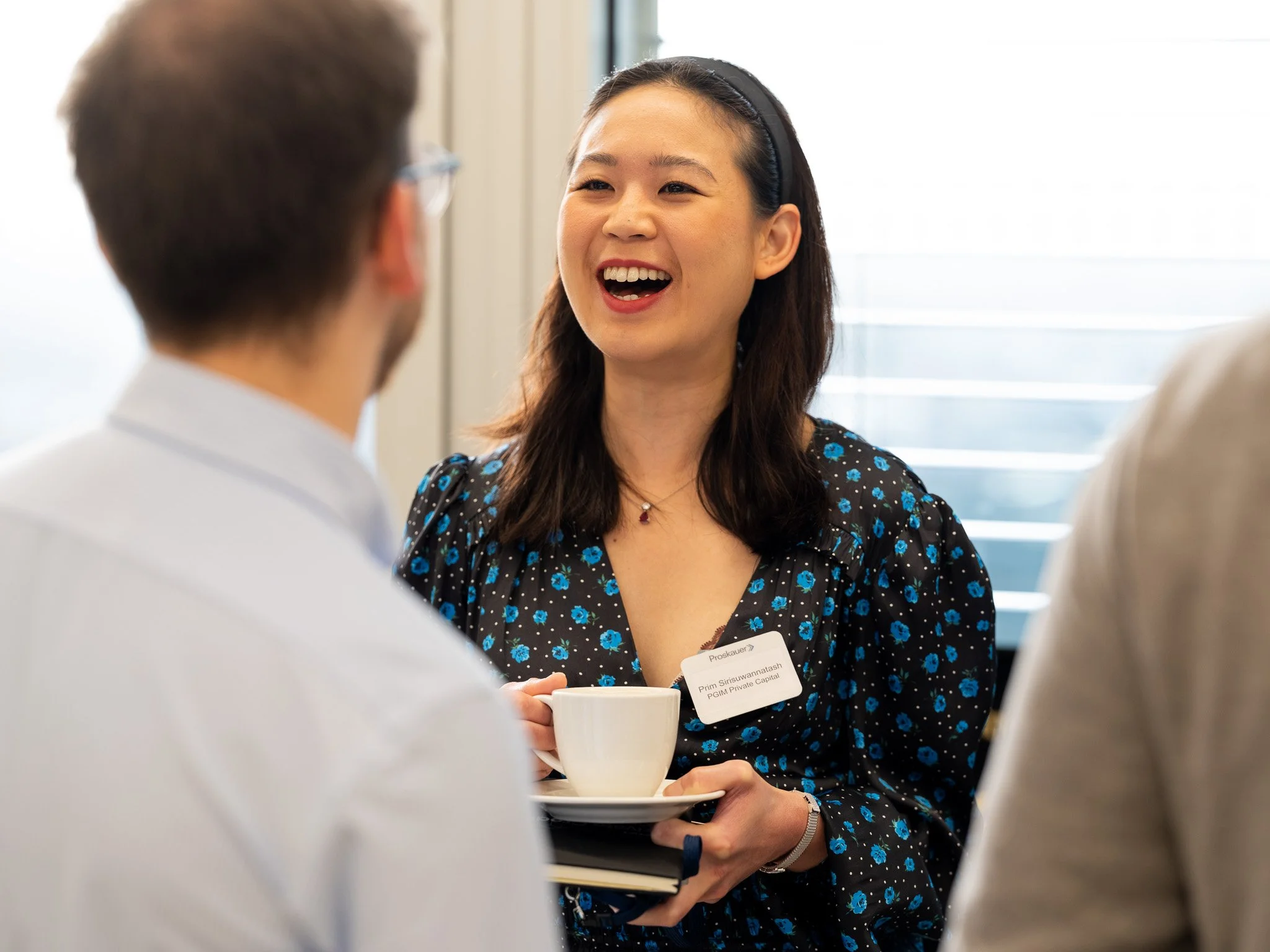 A woman with dark hair wearing a black floral dress and a name badge is talking to two men at a professional event, holding a white teacup and saucer.