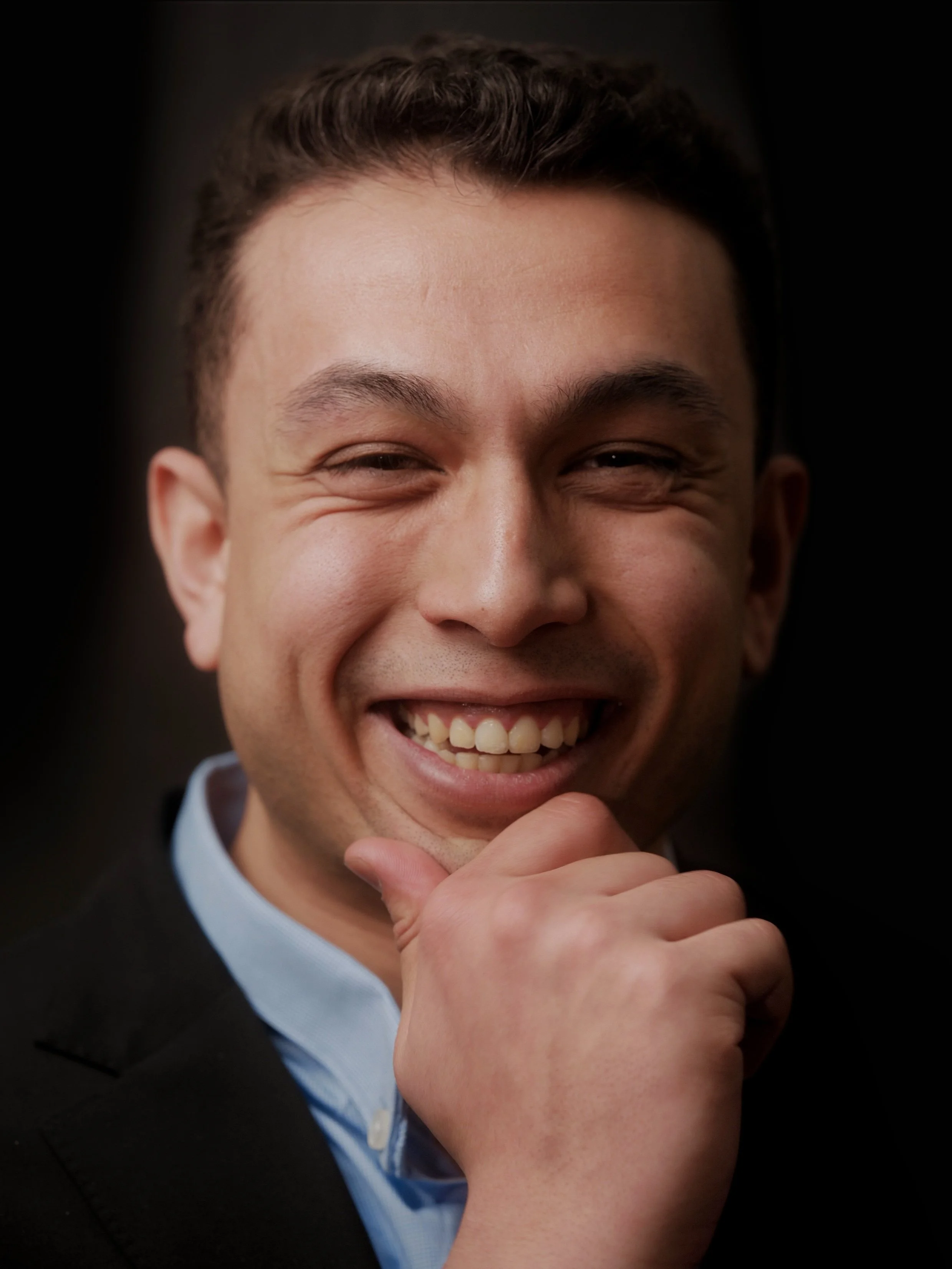 Close-up of a smiling man with short dark hair, wearing a black suit and light blue shirt, with his hand resting on his chin.