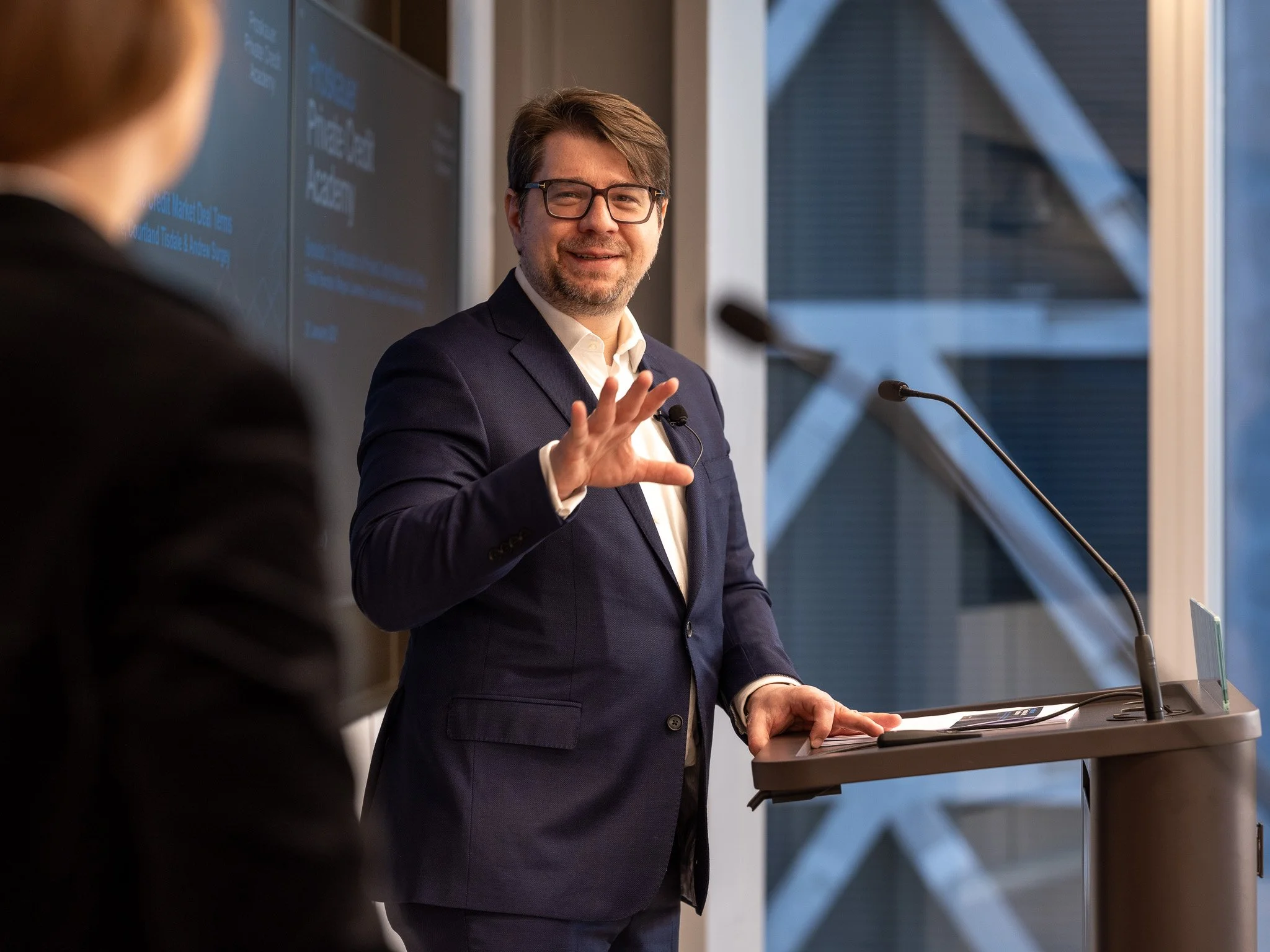 A man in a suit and glasses giving a presentation in a modern conference room, with a woman in the foreground.