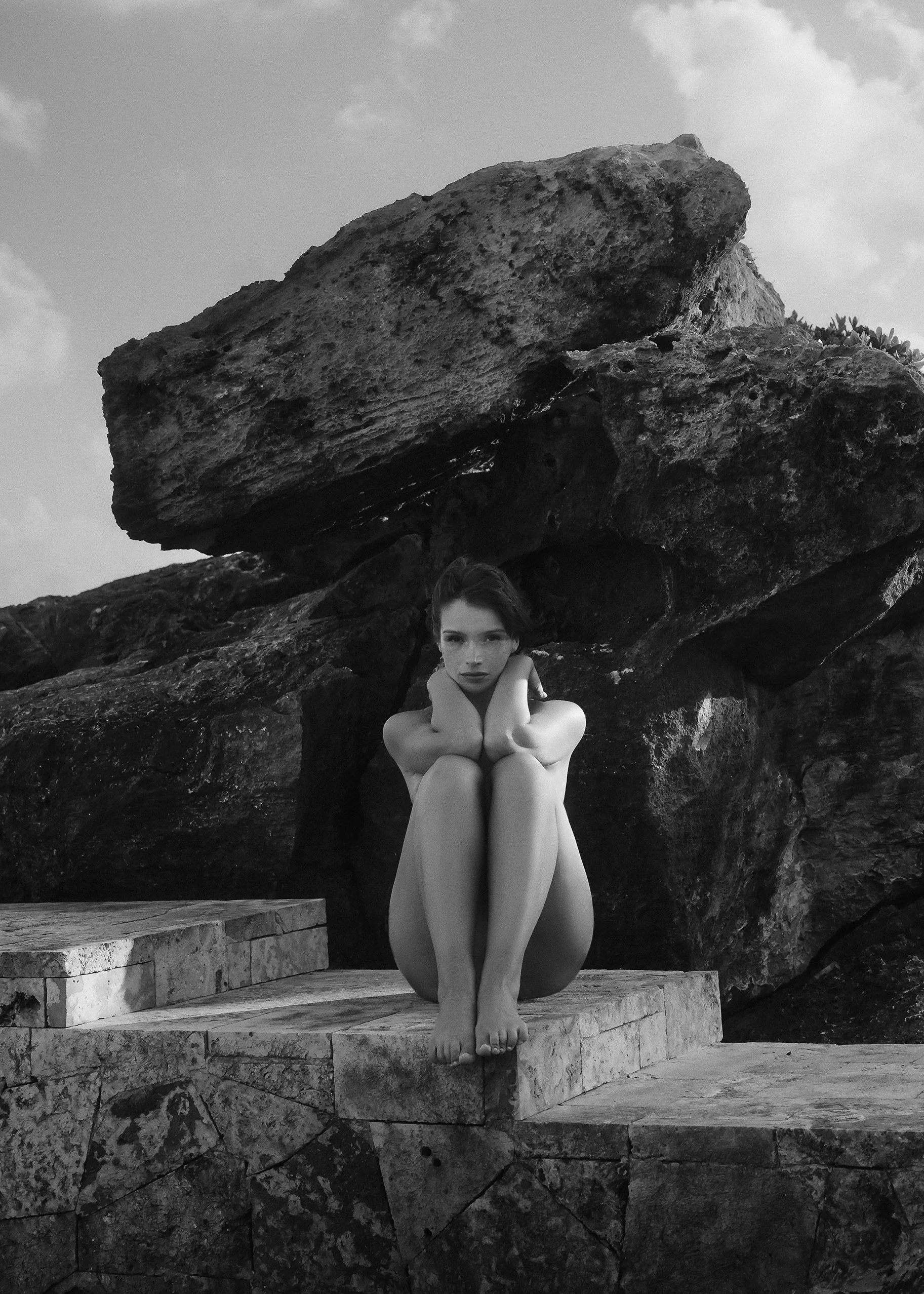 Black and white portrait of a woman seated on stone steps in front of a massive rock formation.
