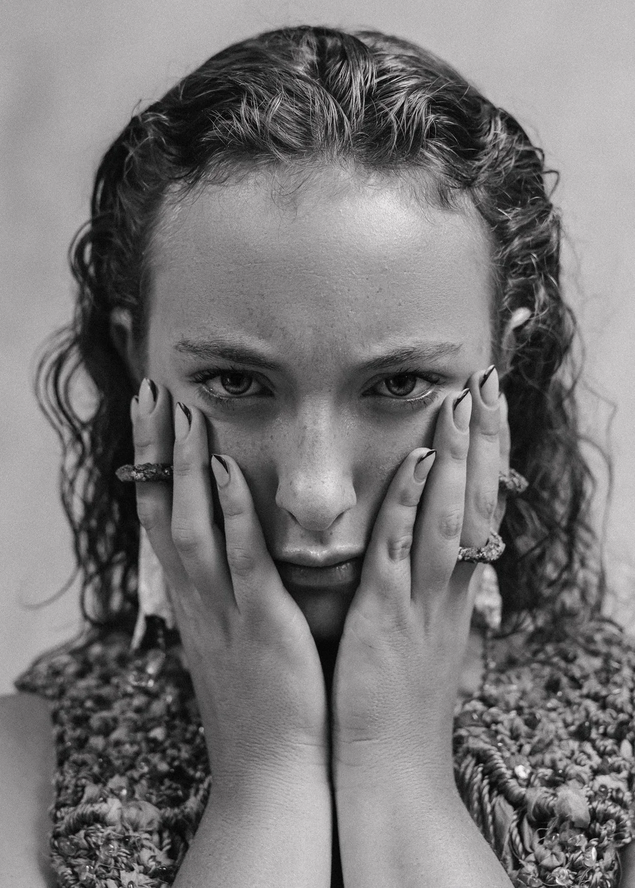Retrato en blanco y negro de una mujer con cabello mojado sosteniendo su rostro con las manos, usando anillos texturizados.
