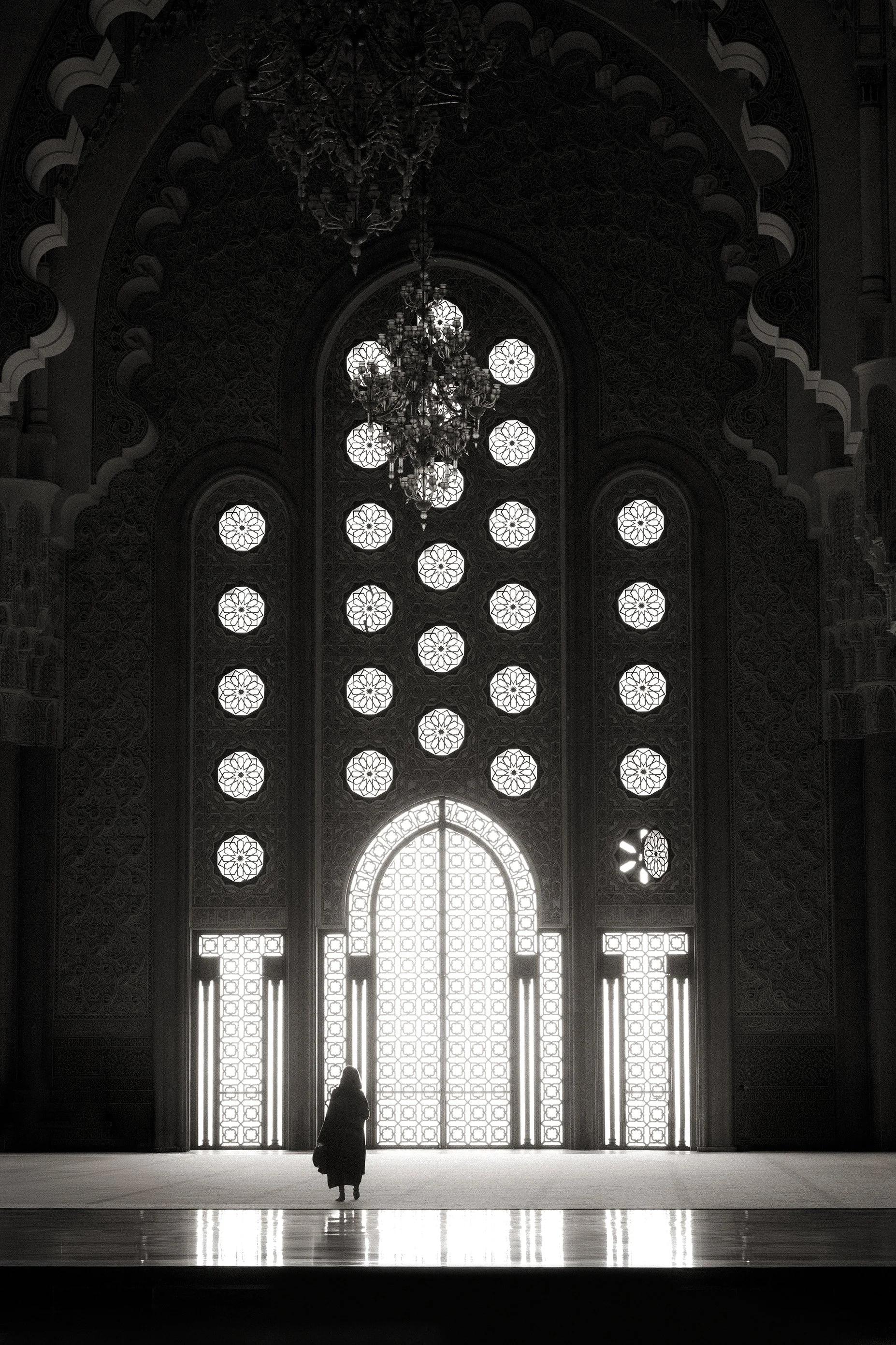 Fine art photograph of a solitary silhouette standing before a massive, ornately patterned window, capturing a forced pause of absolute presence within monumental space.