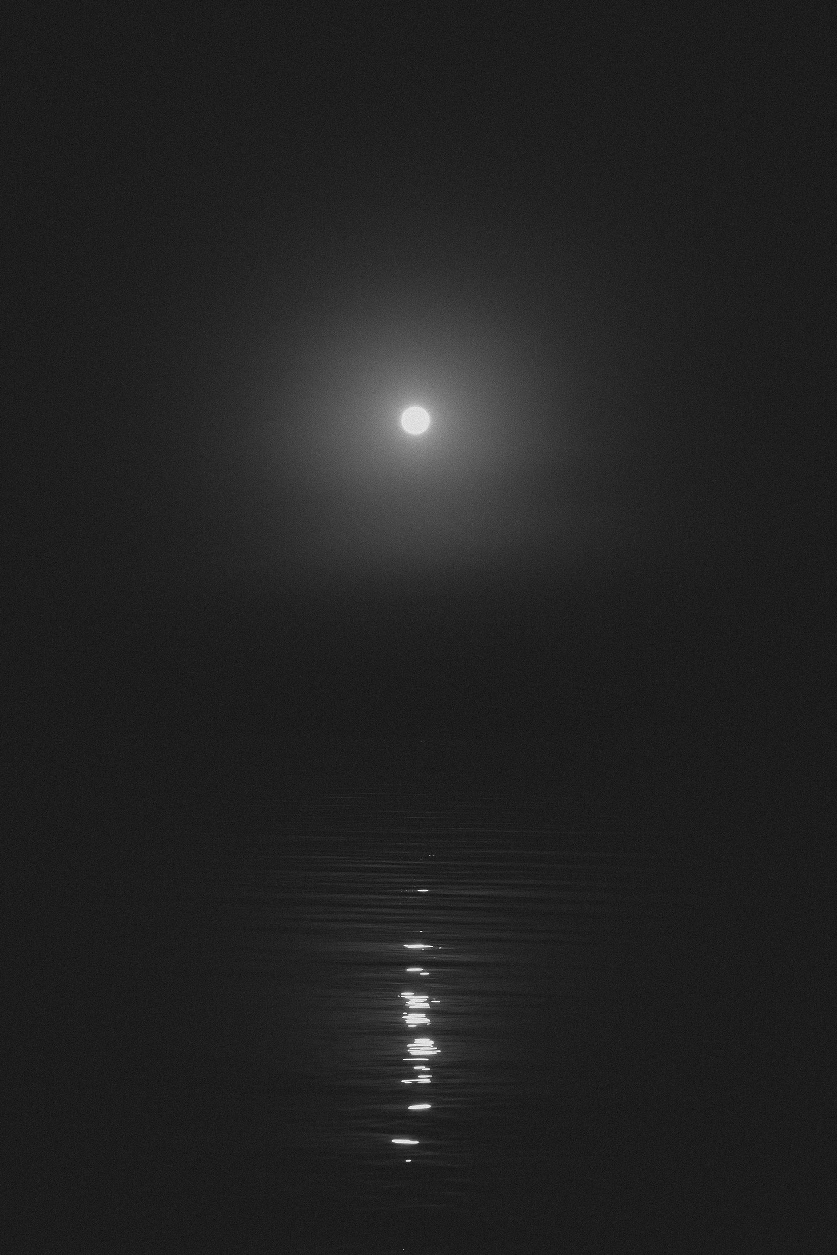 Fotografía en blanco y negro de la luna llena reflejándose sobre el mar en la oscuridad.