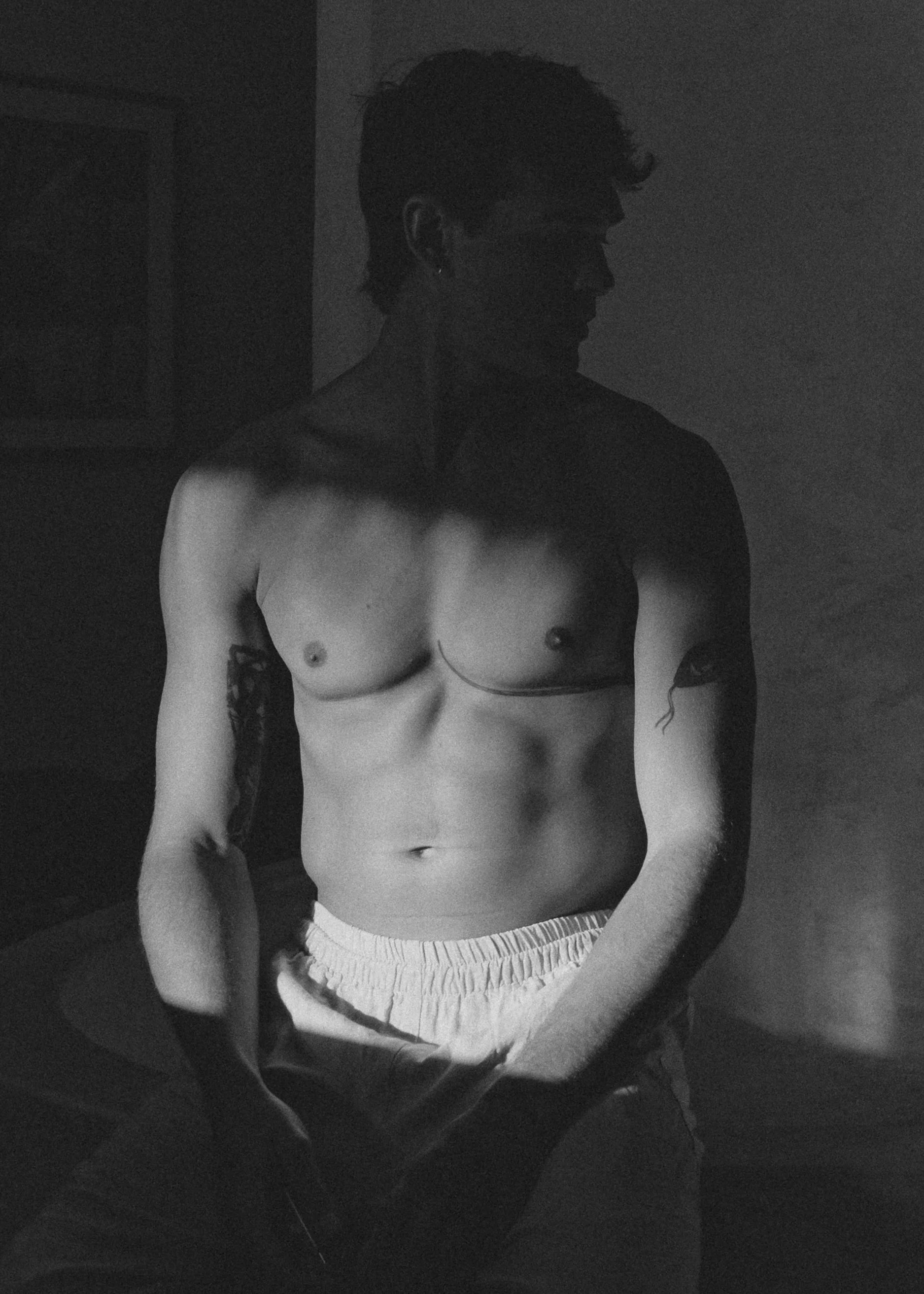 Black and white studio portrait of a shirtless man seated, crossed by a harsh stripe of light and shadow across his torso.