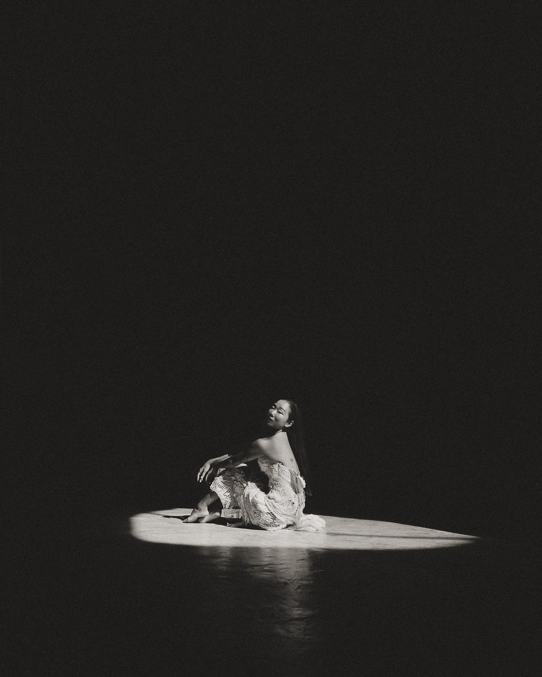 Fotografía en blanco y negro de una mujer sentada sobre una superficie iluminada por la luz de una ventana en medio de la oscuridad.