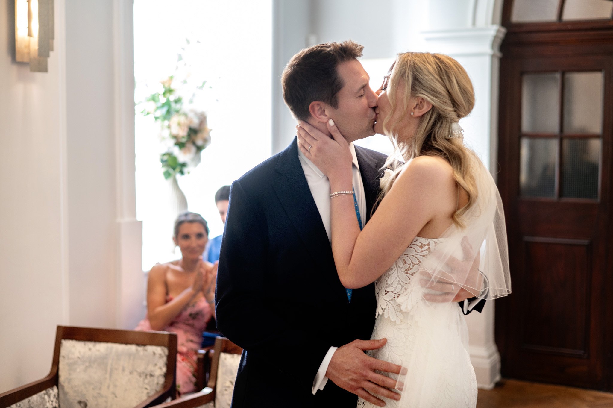 A bride and groom kiss during their wedding ceremony with three guests clapping in the background.
