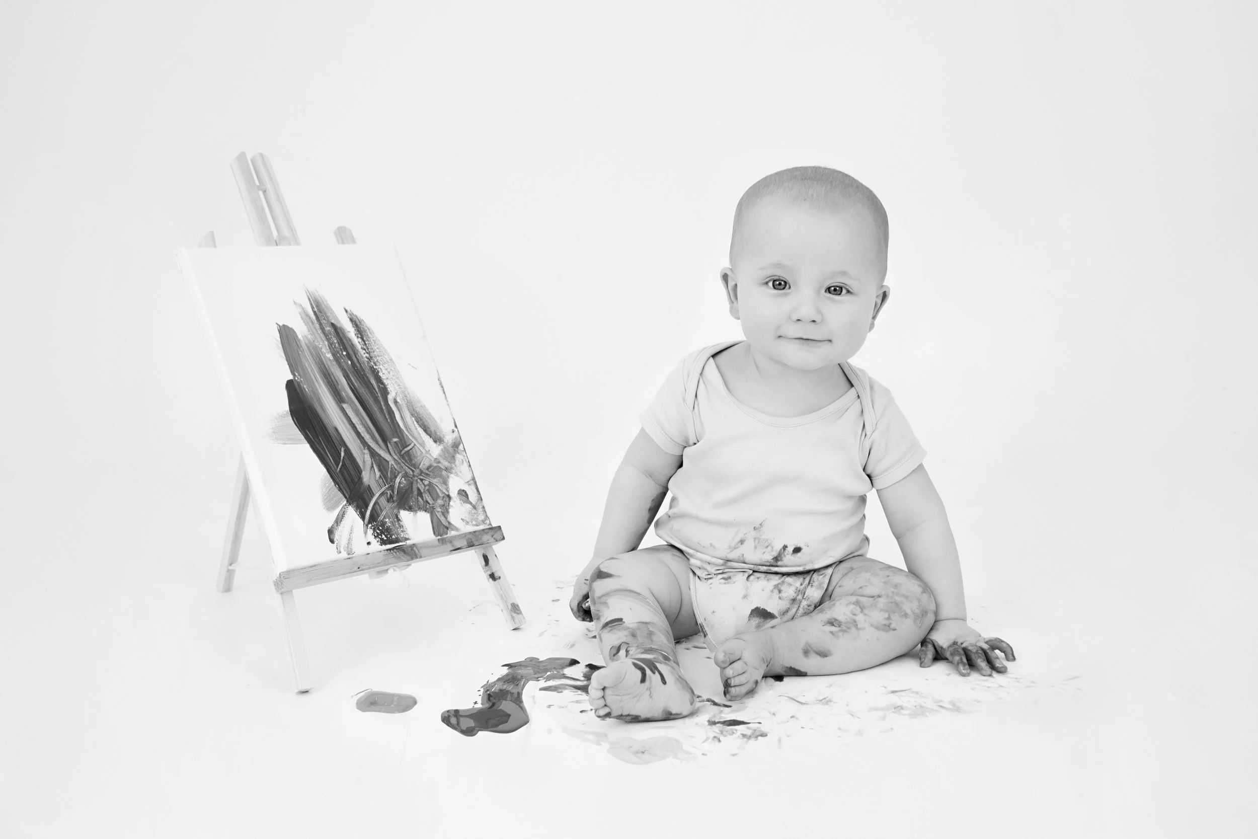 Black and white photo of a baby sitting on the floor covered in paint, next to a canvas with abstract brush strokes and paint splatters.