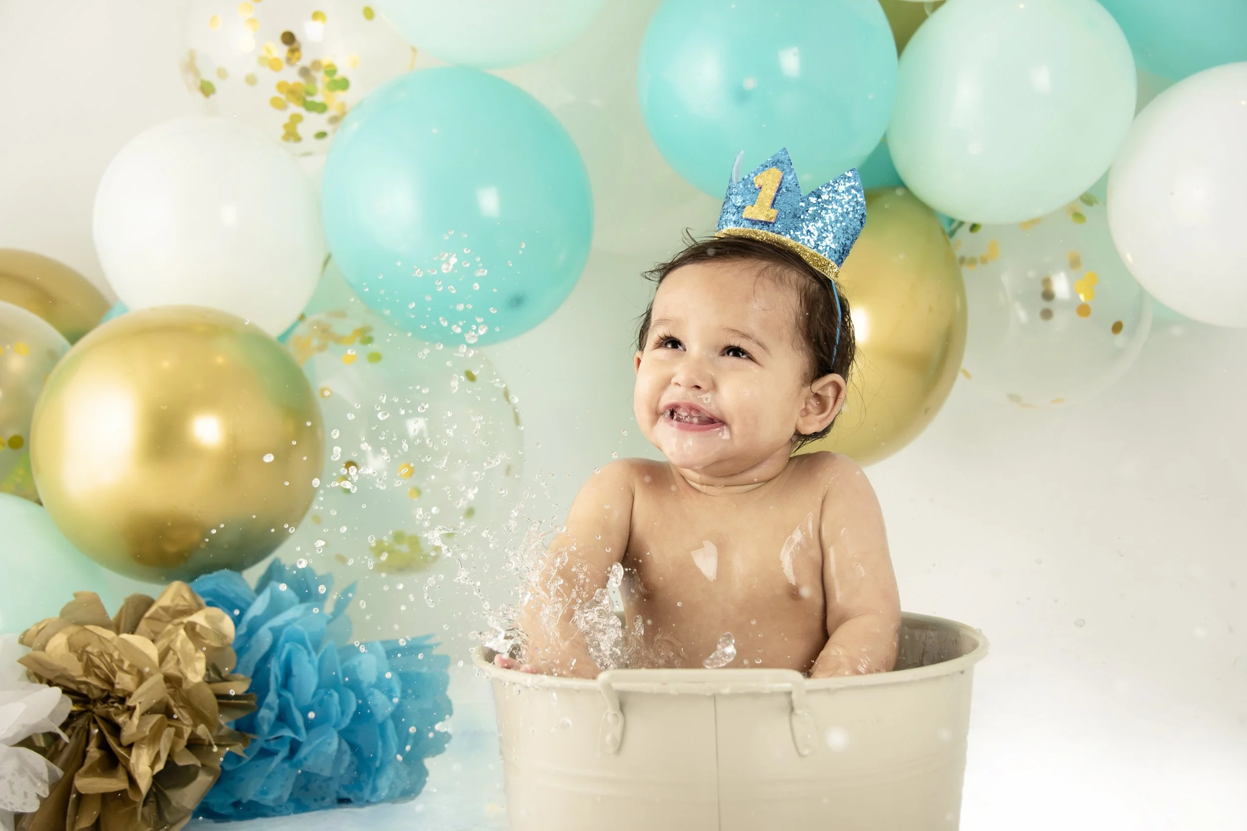 Baby in a tub with blue birthday crown surrounded by colorful balloons and decorations.