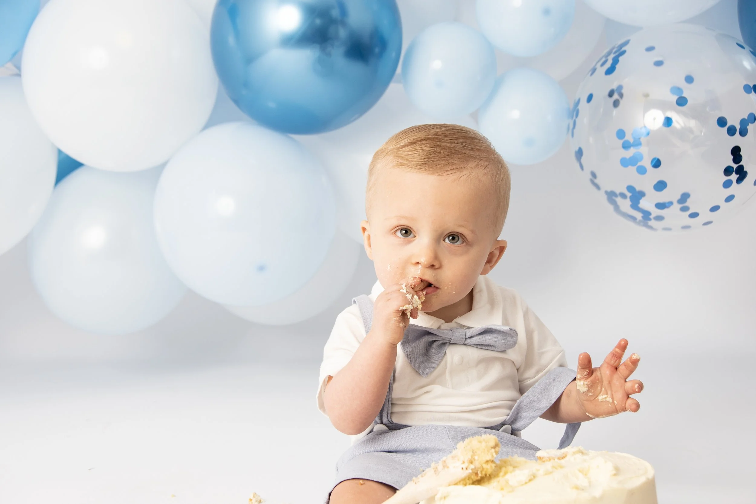 Baby eating cake with blue and white balloons in the background.