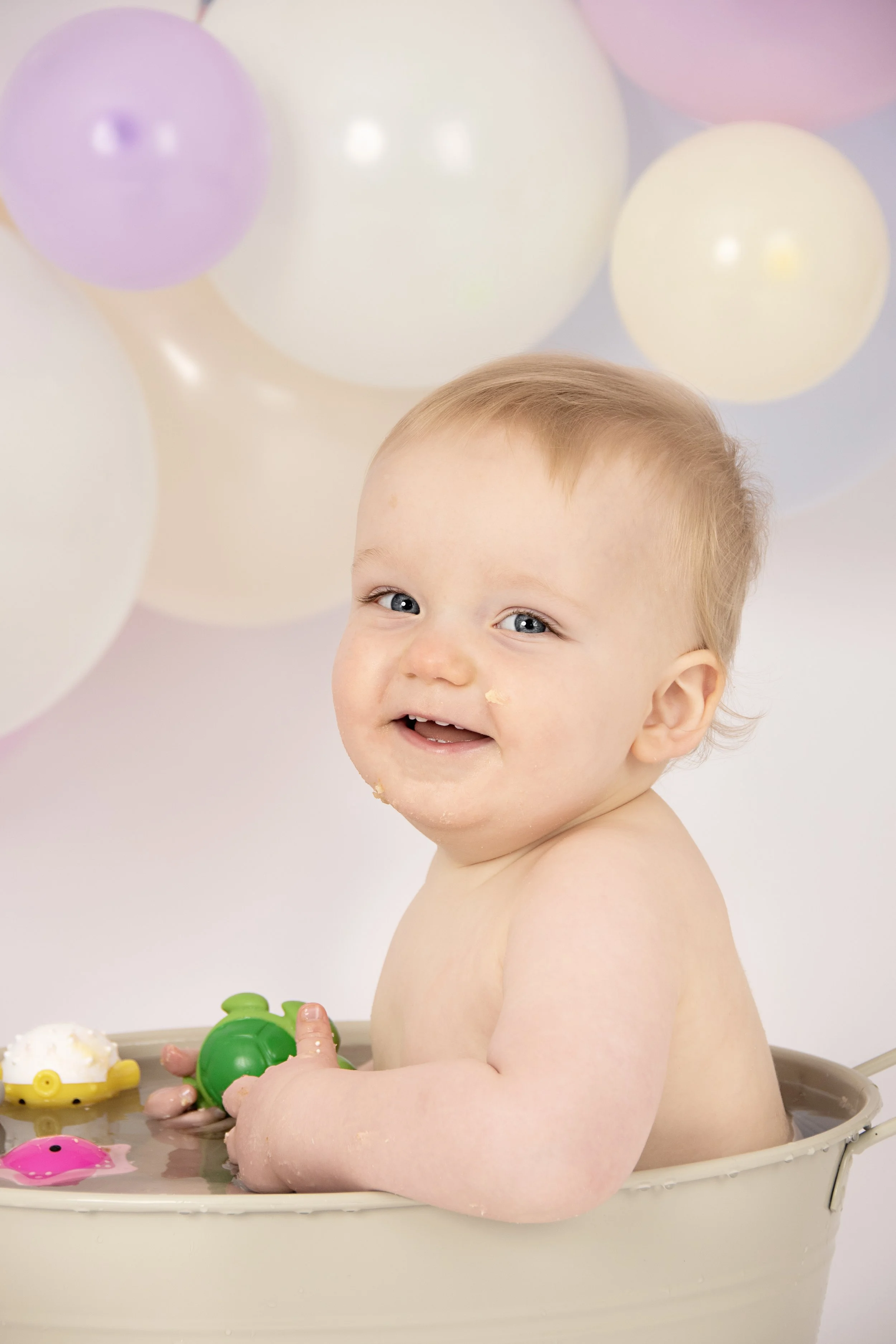Smiling baby sitting in a tub with colorful balloons in the background.