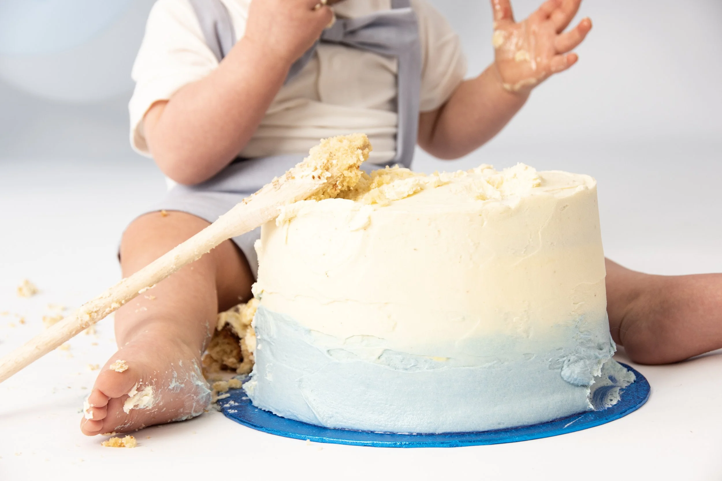 Baby in diaper sitting with a large cream and blue cake, using a spatula to dig in and get messy, hands and feet covered in frosting.