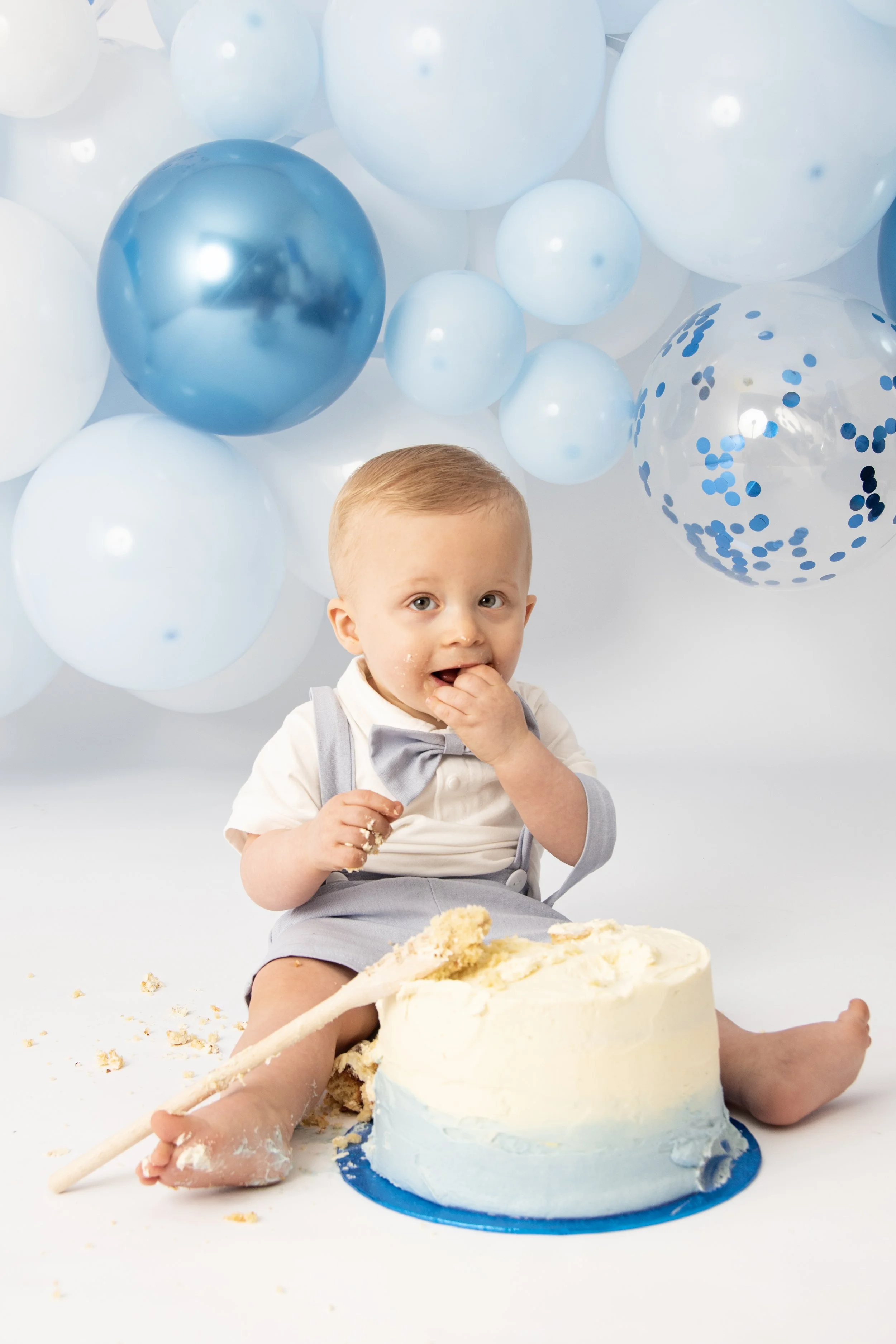 A baby wearing a blue bow tie and suspenders sits on the floor, eating cake with their hands. There is a blue and white layered cake in front of them, partially smashed. The background features a cluster of blue and white balloons.