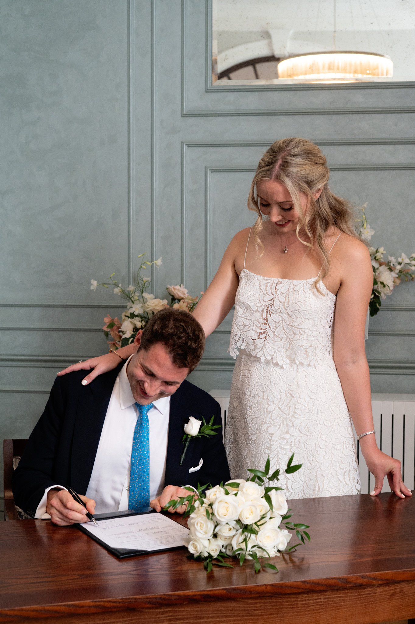 A woman in a white lace dress places her hand on a man's shoulder who is signing a document at a wedding. A bouquet of white roses is on the table, with pink flowers behind them.