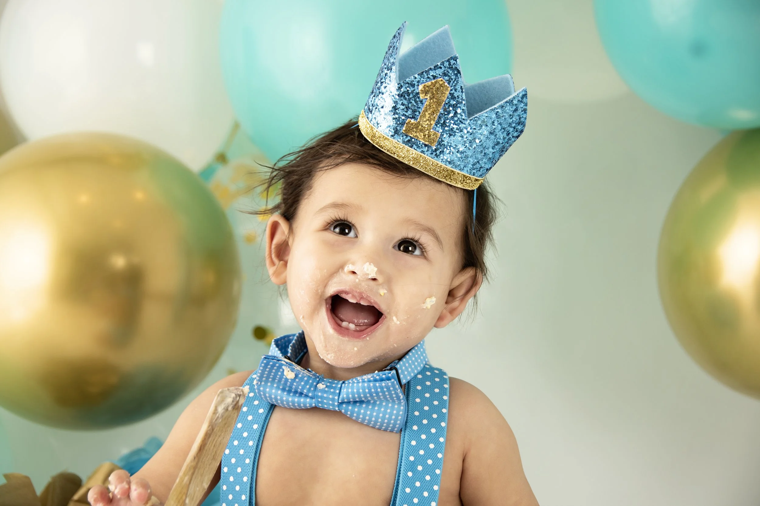 Smiling baby wearing a blue glitter crown with a gold number one, polka dot blue bow tie and suspenders, surrounded by large gold and teal balloons, icing on face.