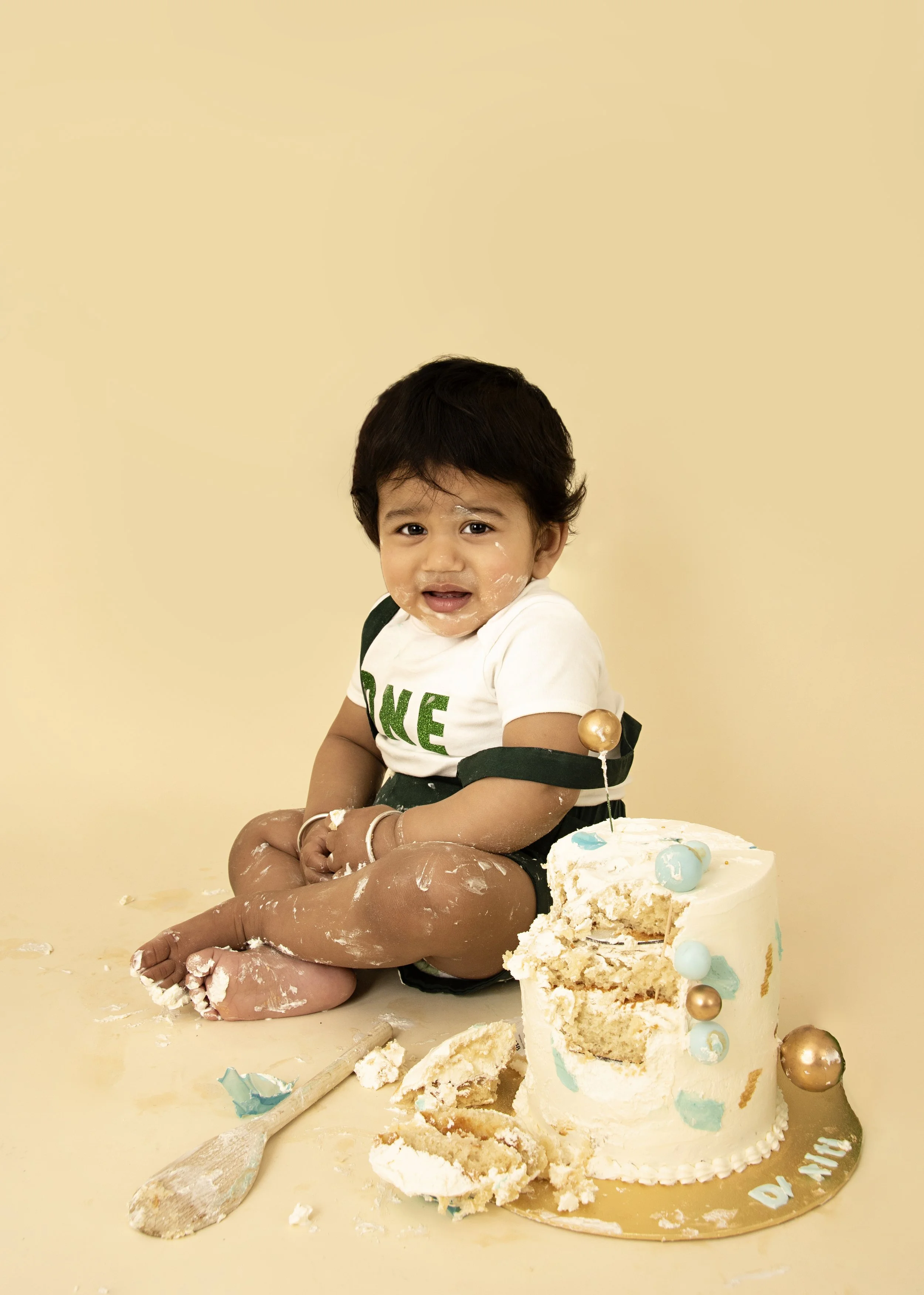 Baby sitting next to a partially eaten cake with icing on their face and hands, wearing a "one" shirt, against a beige background.