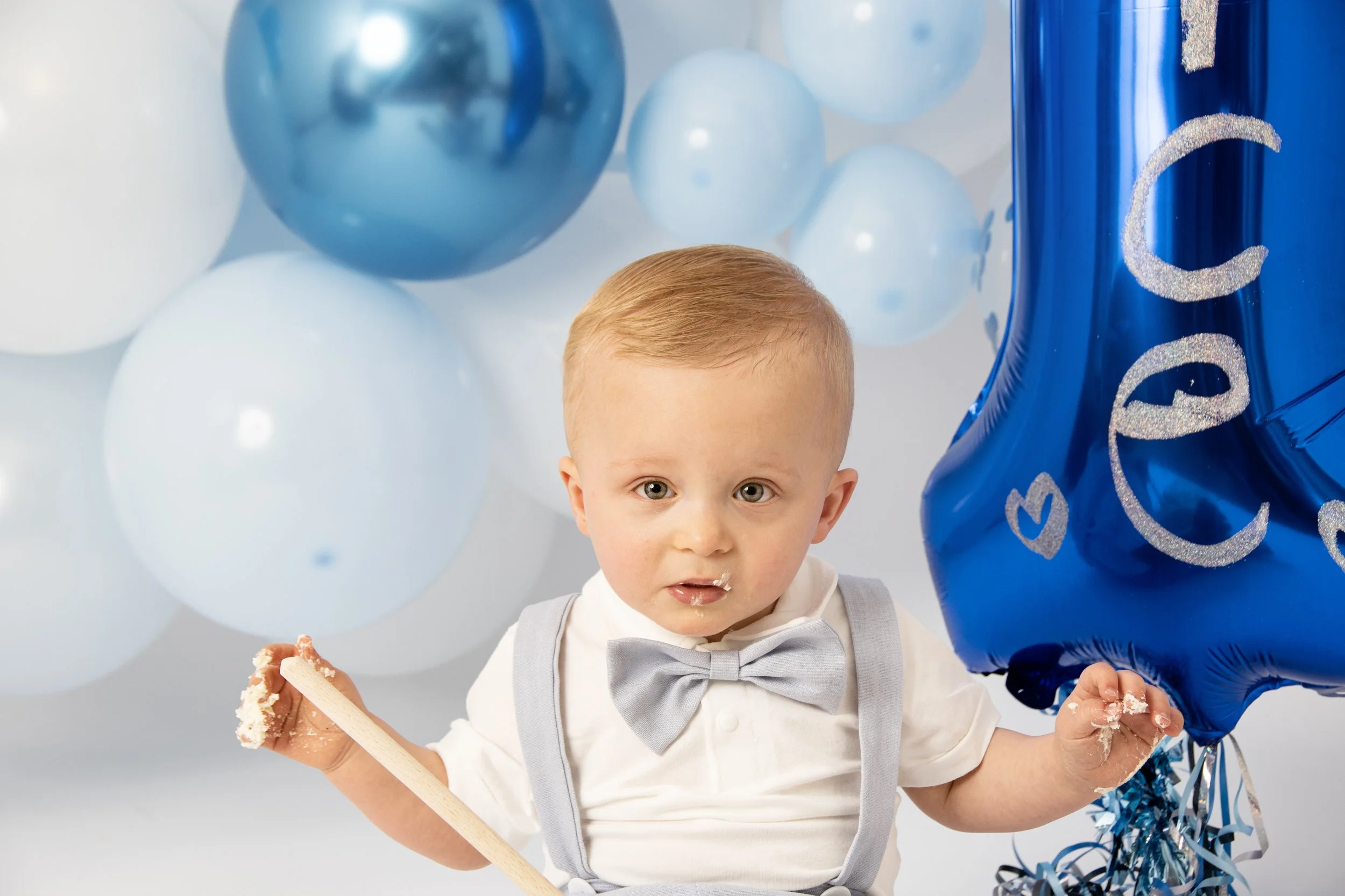 A toddler wearing a bow tie and suspenders with cake on face and hands, surrounded by blue and white balloons, holding a blue letter-shaped balloon.