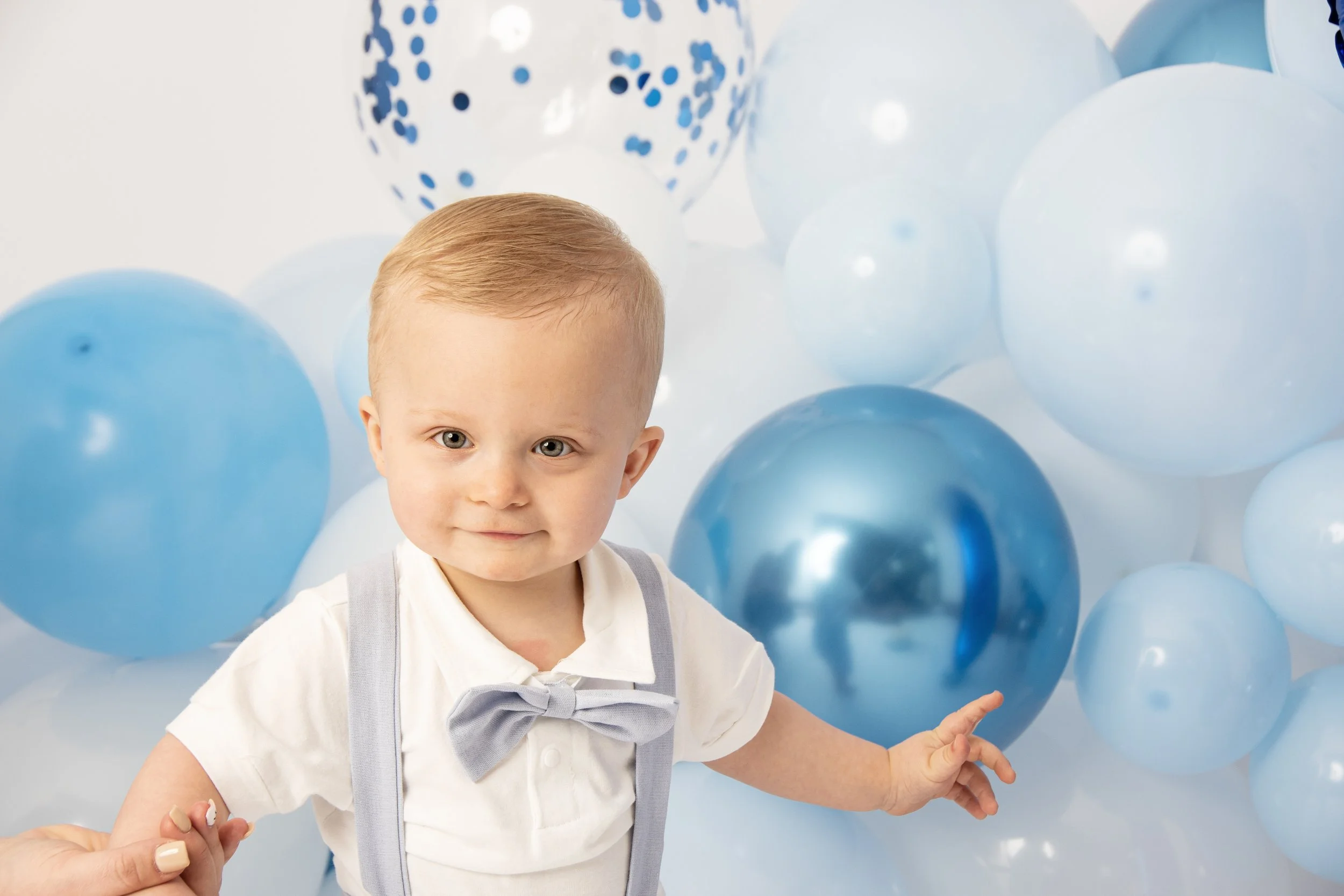 Toddler in white shirt and bow tie with blue backdrop of balloons