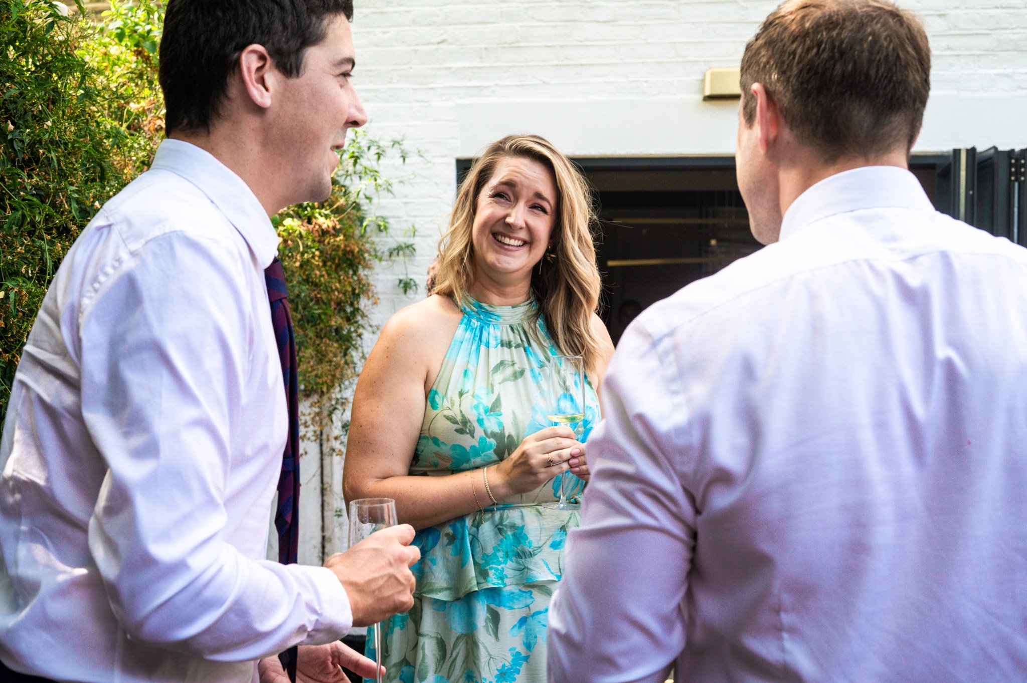 A woman in a floral dress laughing and holding a wine glass, talking to two men in white shirts at an outdoor gathering.
