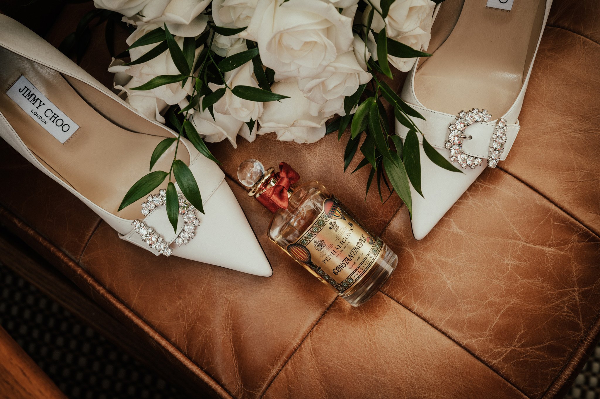 Pair of white Jimmy Choo high heels with jewel embellishments on the toes, surrounded by white flowers and green leaves, with a bottle of Penhaligon's perfume on a brown leather surface.