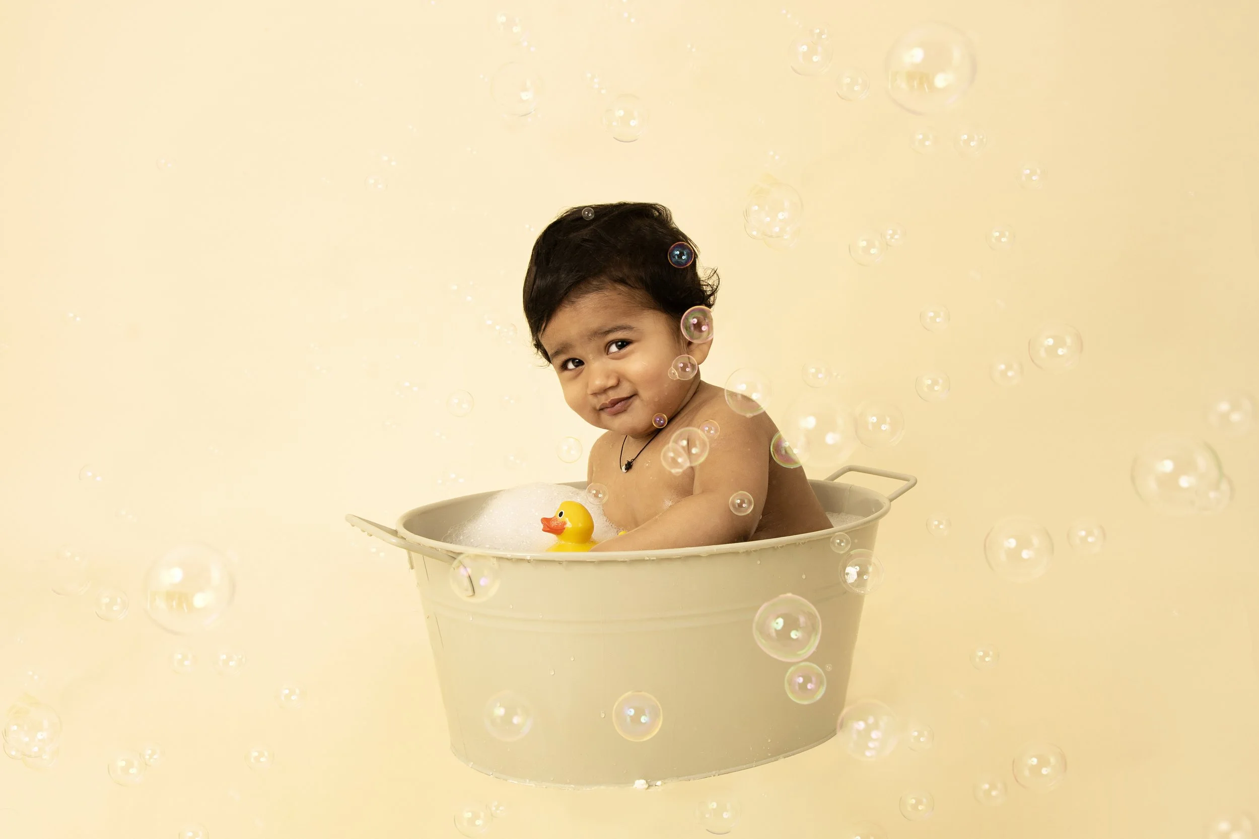 A baby sitting in a metal tub filled with bubbles and a rubber duck surrounded by floating bubbles.