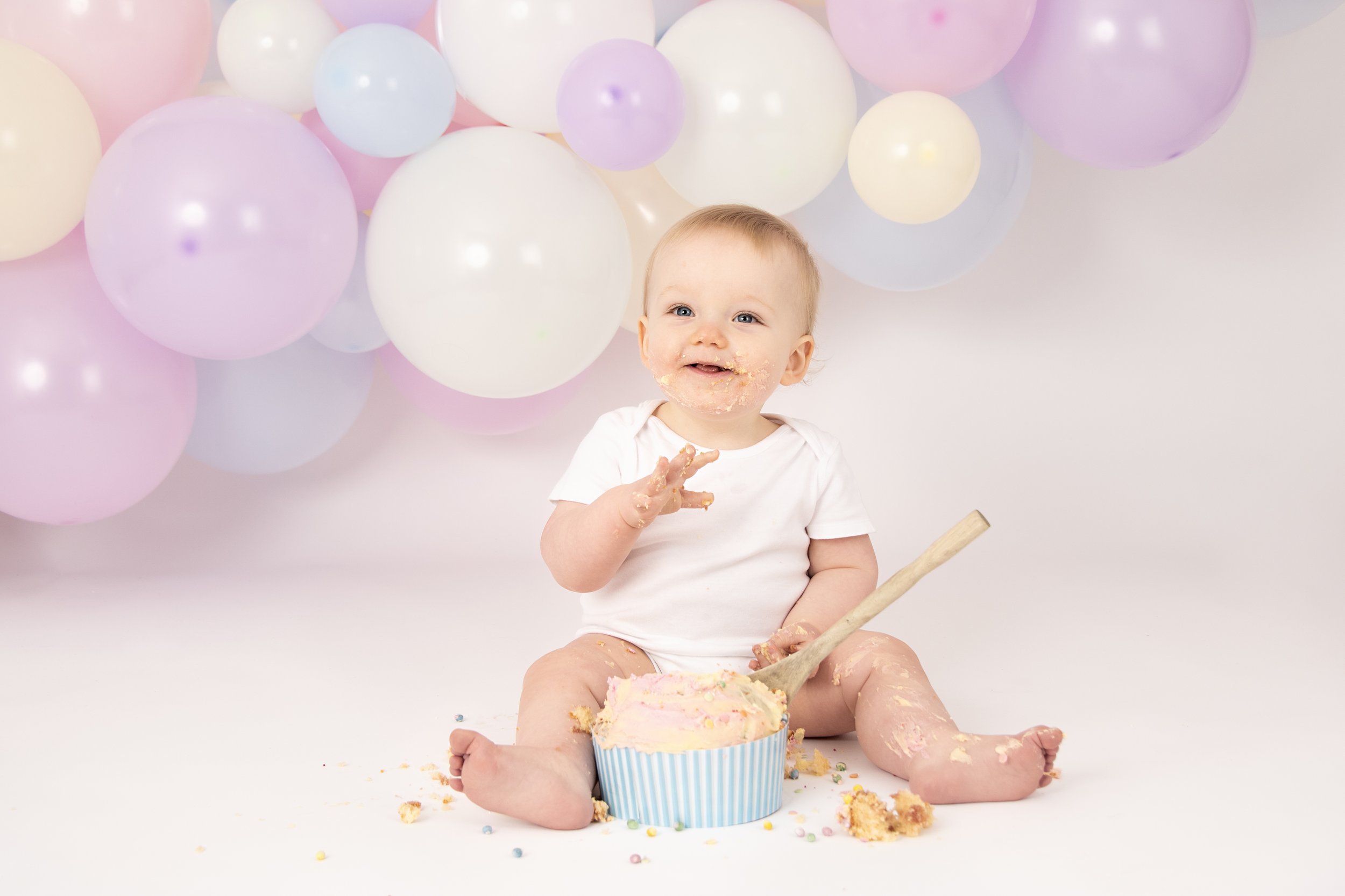 Baby sitting on floor with cake and balloons, smiling and covered in frosting.