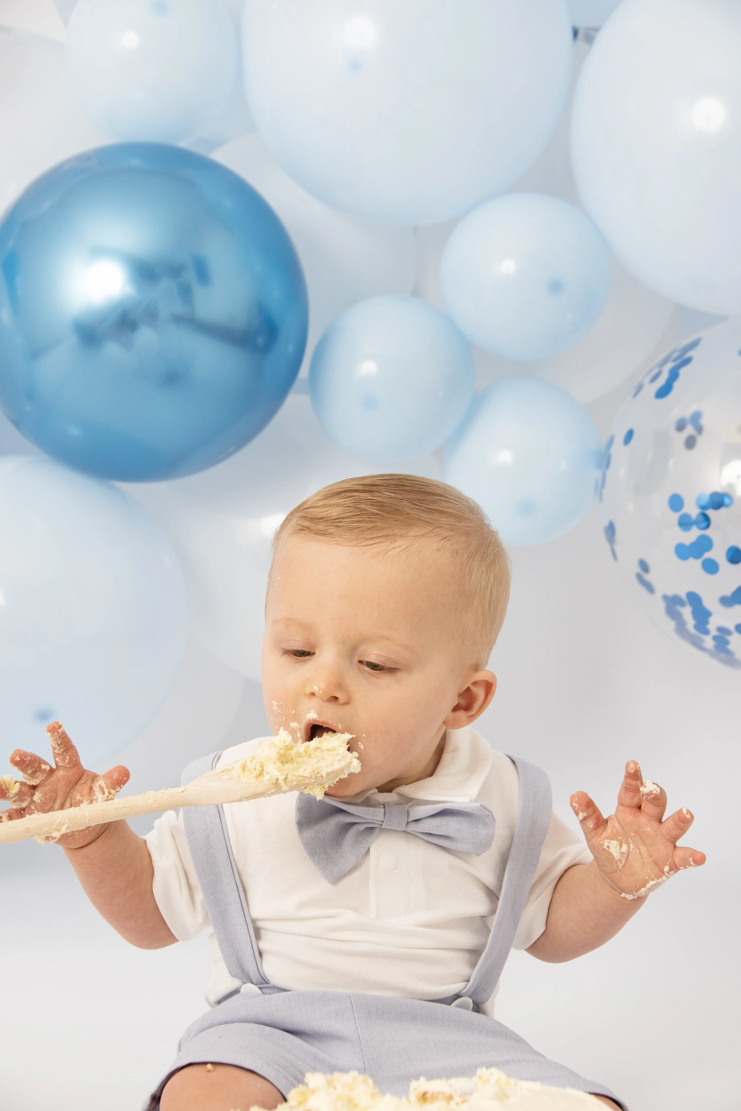 Baby in light blue outfit eating cake with a spoon, surrounded by blue and white balloons.