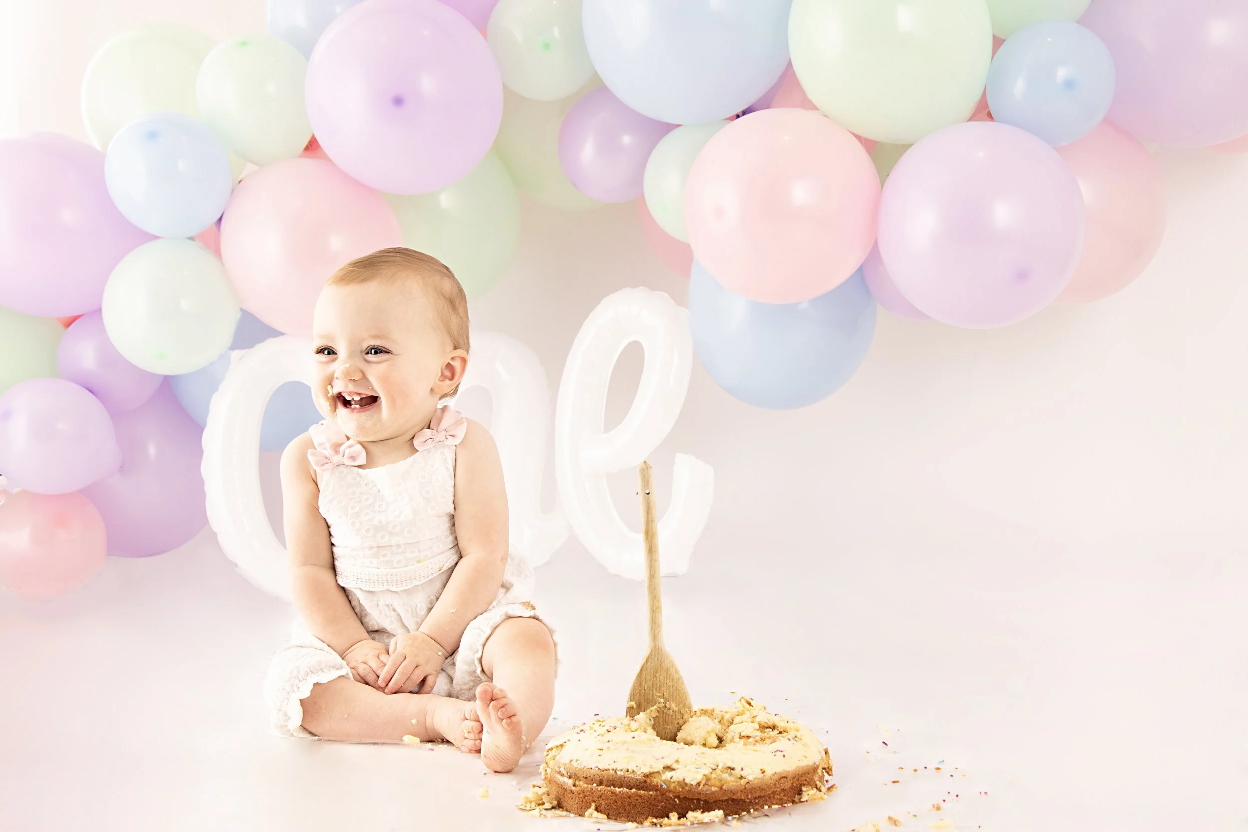 Smiling baby sitting on floor with cake, pastel balloons in background.